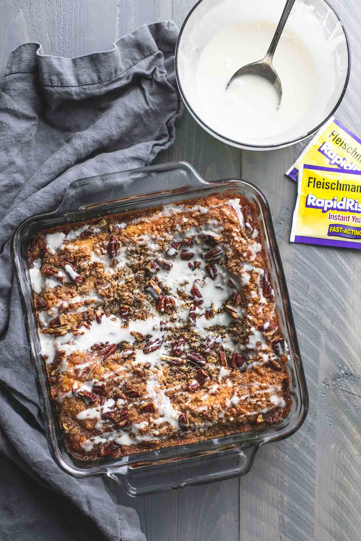 top view of cinnamon pecan coffee cake in a square pan with batter and a spoon in a bowl and rapid rise yeast packets on the side.