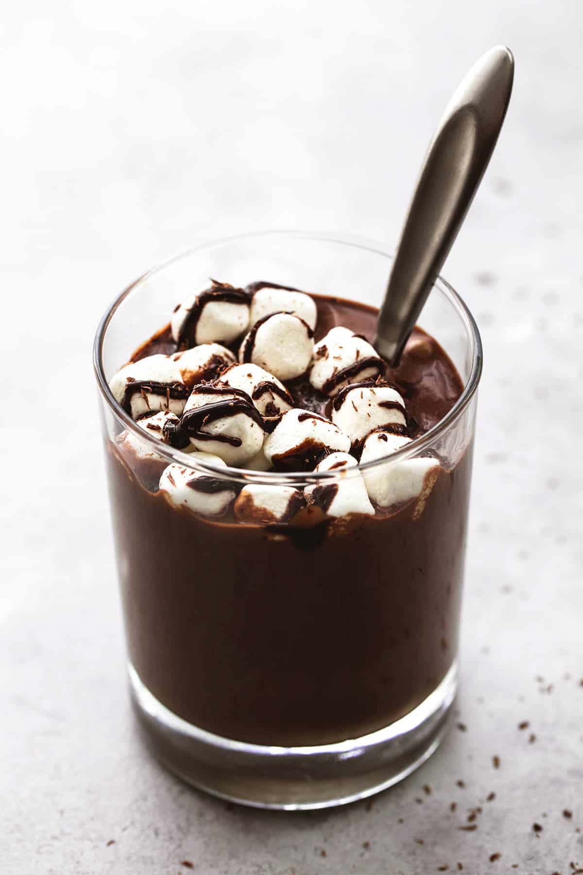 extra thick hot chocolate with a spoon in a glass topped with marshmallows and chocolate syrup.