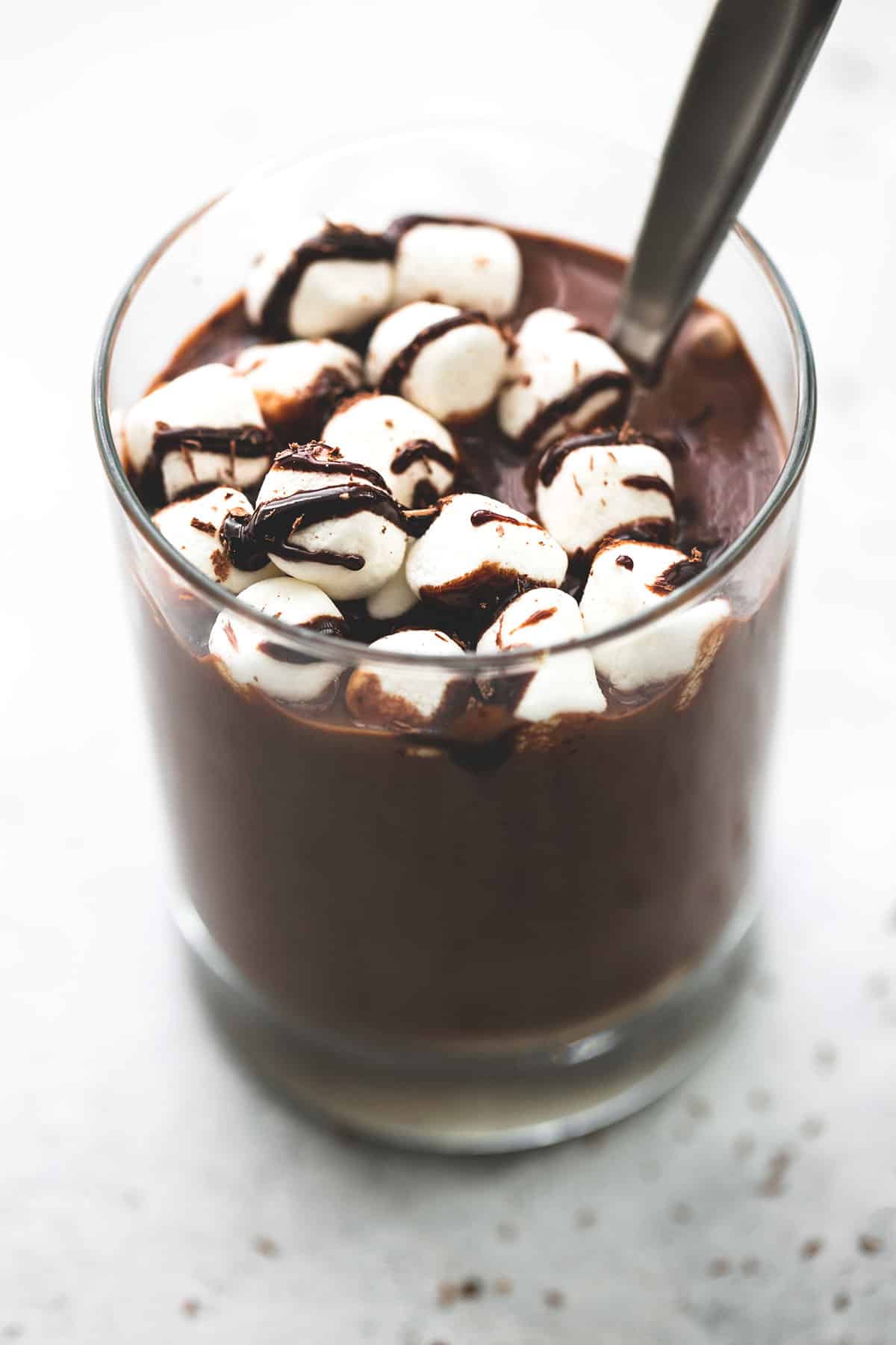 extra thick hot chocolate with a spoon in a glass topped with marshmallows and chocolate syrup.