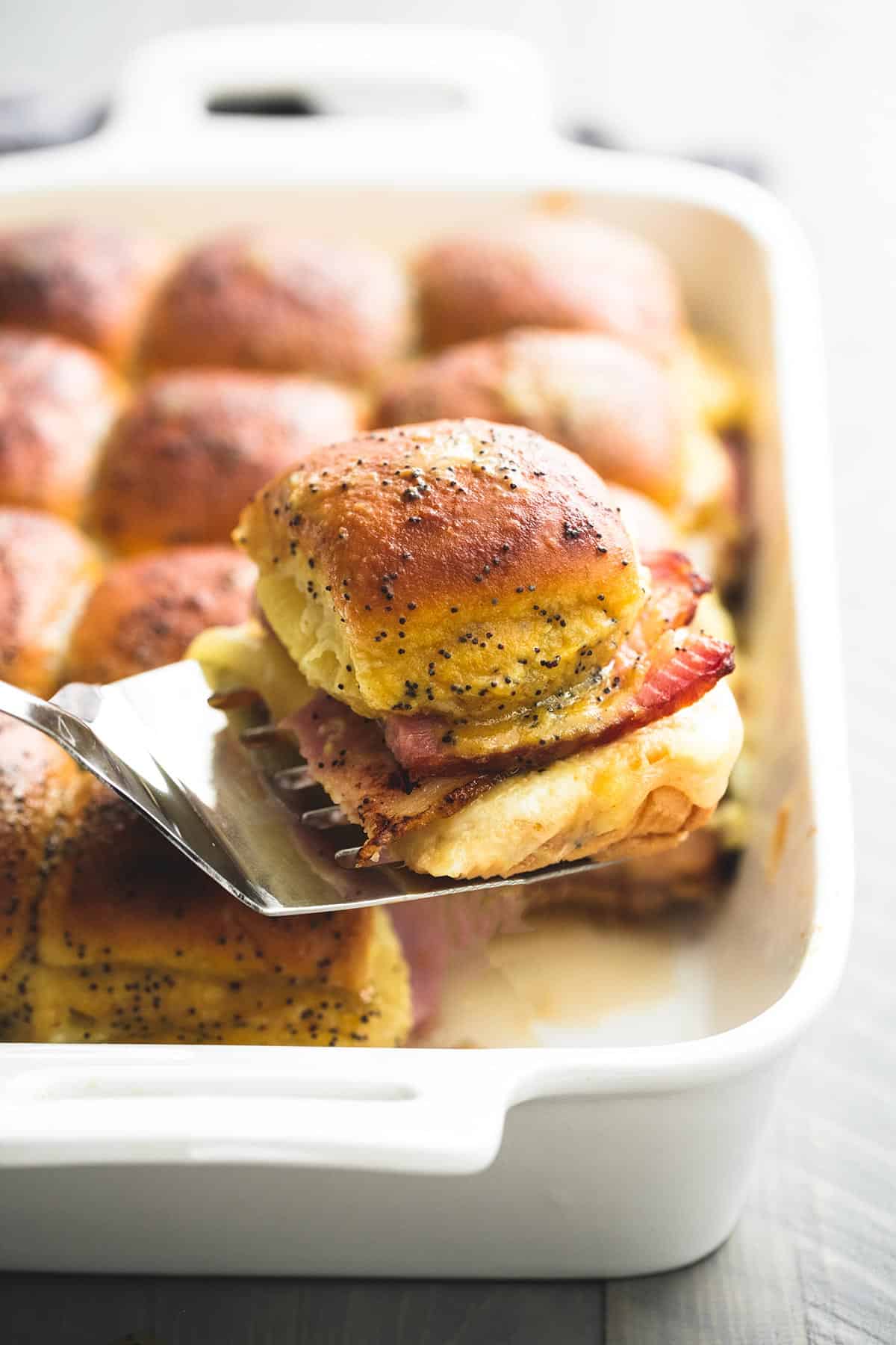 a spatula holding up a ham & swiss cheese slider above more sliders in a baking pan.