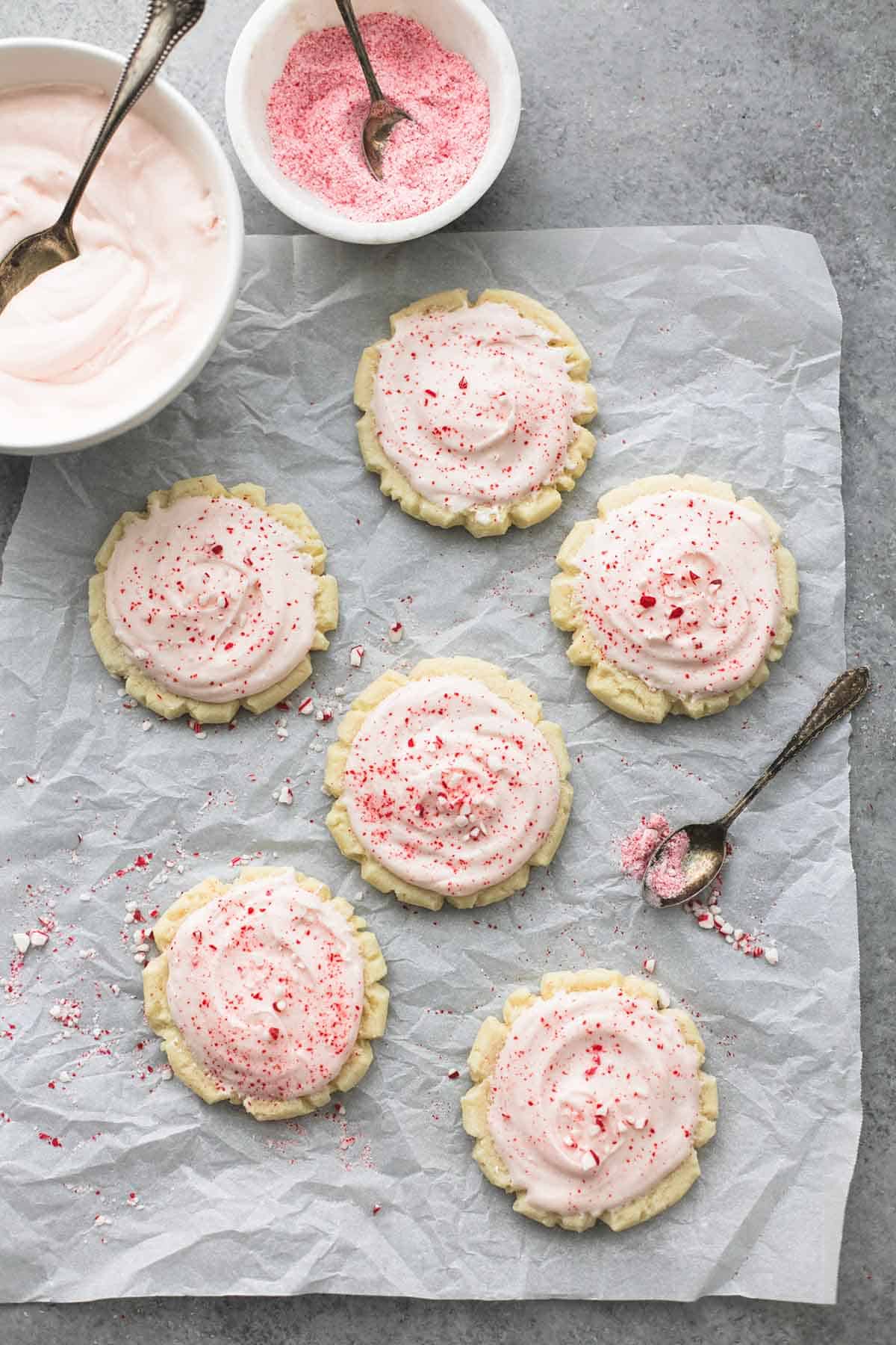 top view of peppermint sugar cookies with a spoon and bowls of frosting and peppermint chunks and spoons on the side.