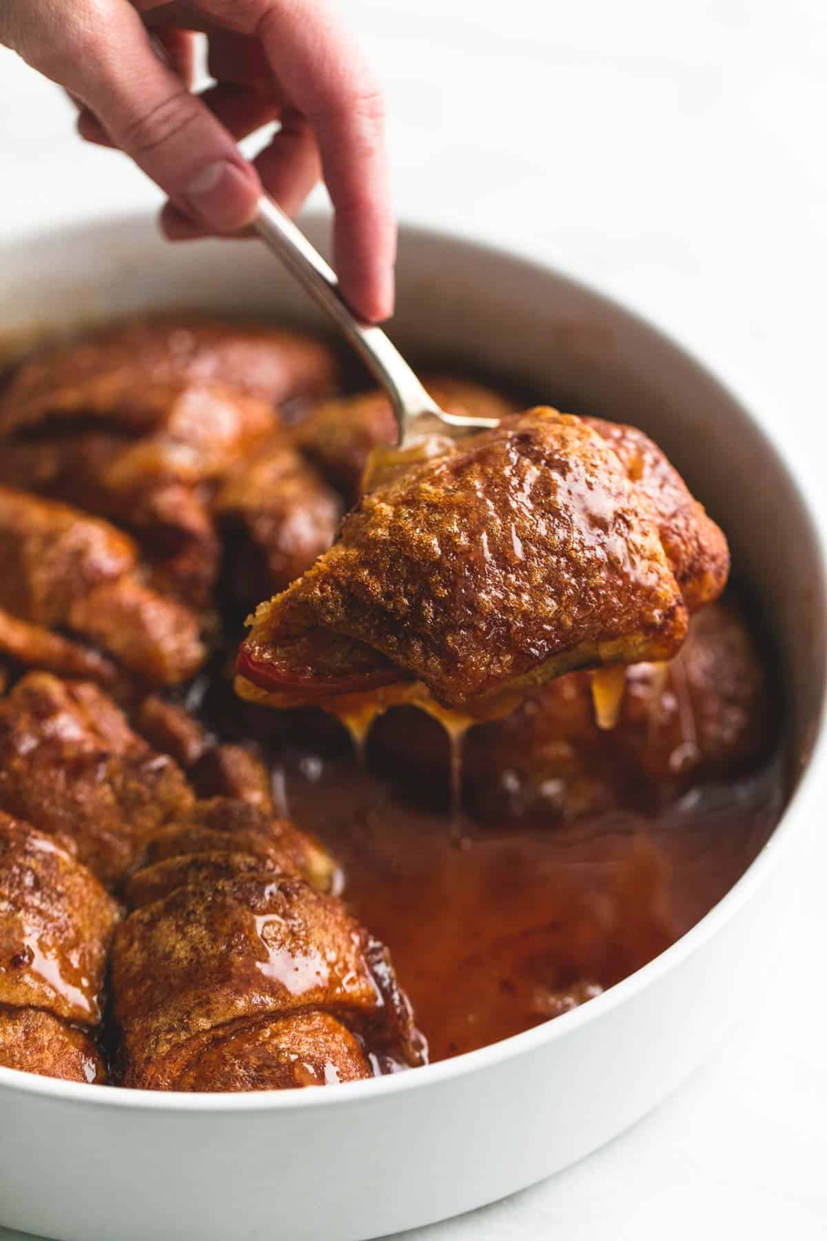 This simple 7UP apple cobbler is made with refrigerated crescent dough and a cinnamon sugar and 7UP caramel sauce that is to die for. | lecremedelacrumb.com a hand holding up a spoon with some apple cobbler on it above a round tray of more cobbler.