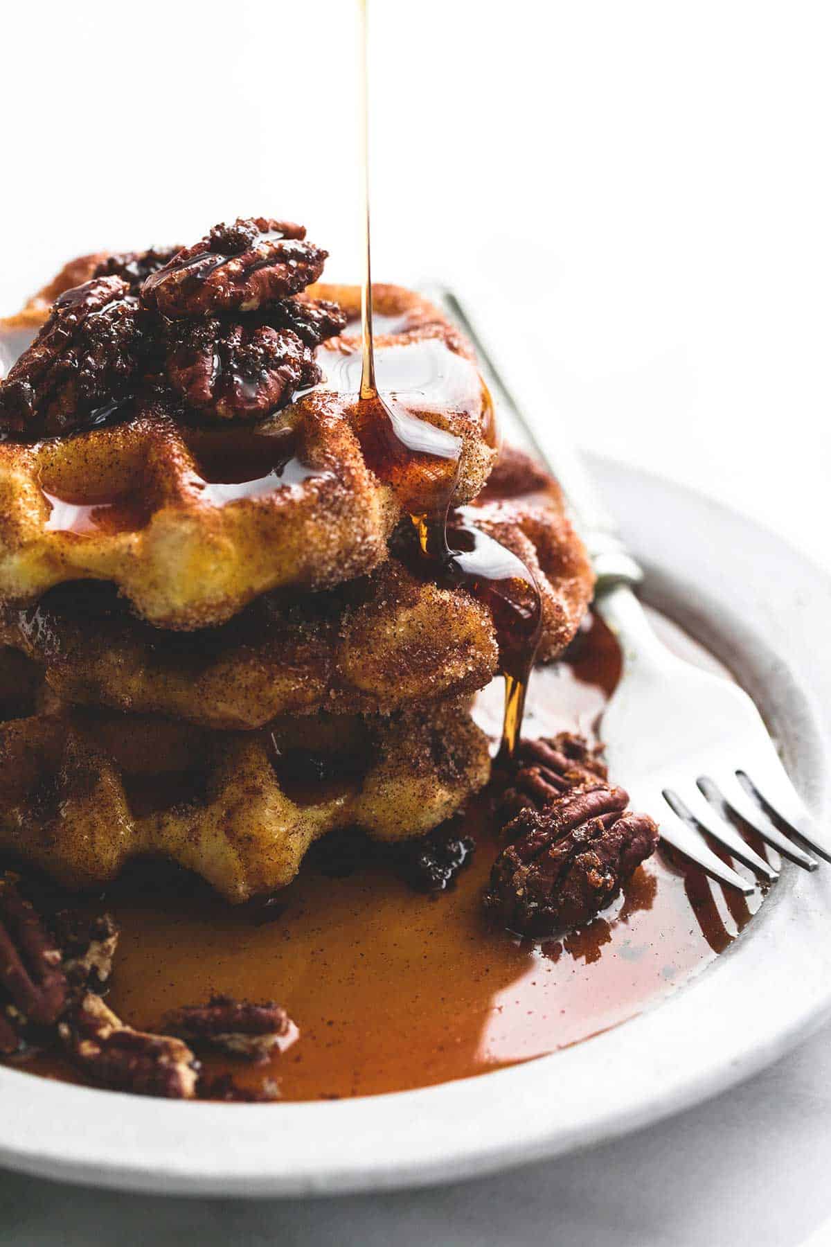 cinnamon sugar biscuit waffles topped with candied pecans with a fork on a plate with syrup being poured on top.