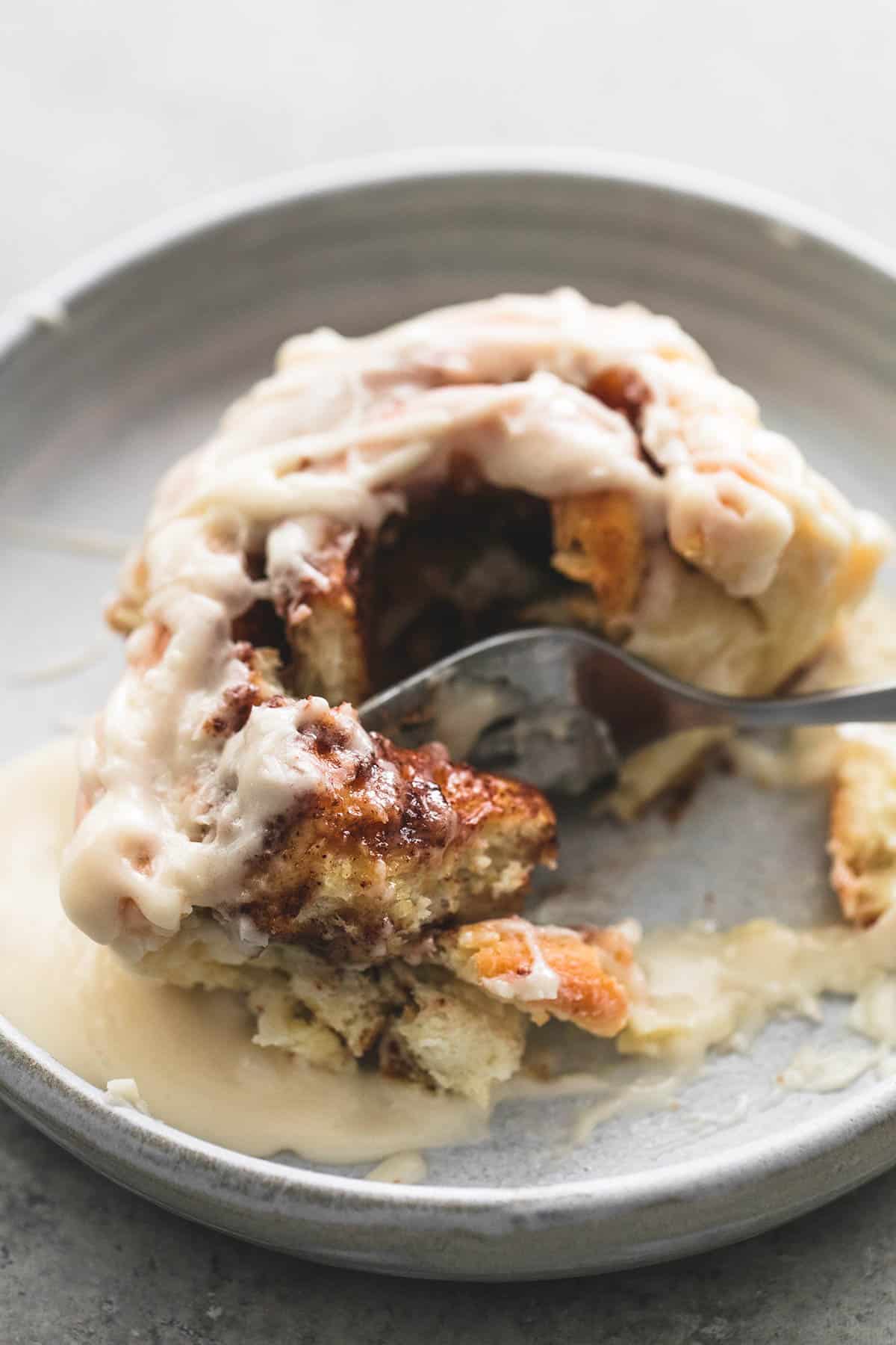 close up of a cream cheese cinnamon roll with a fork in between a bite and the rest of the roll on a plate.