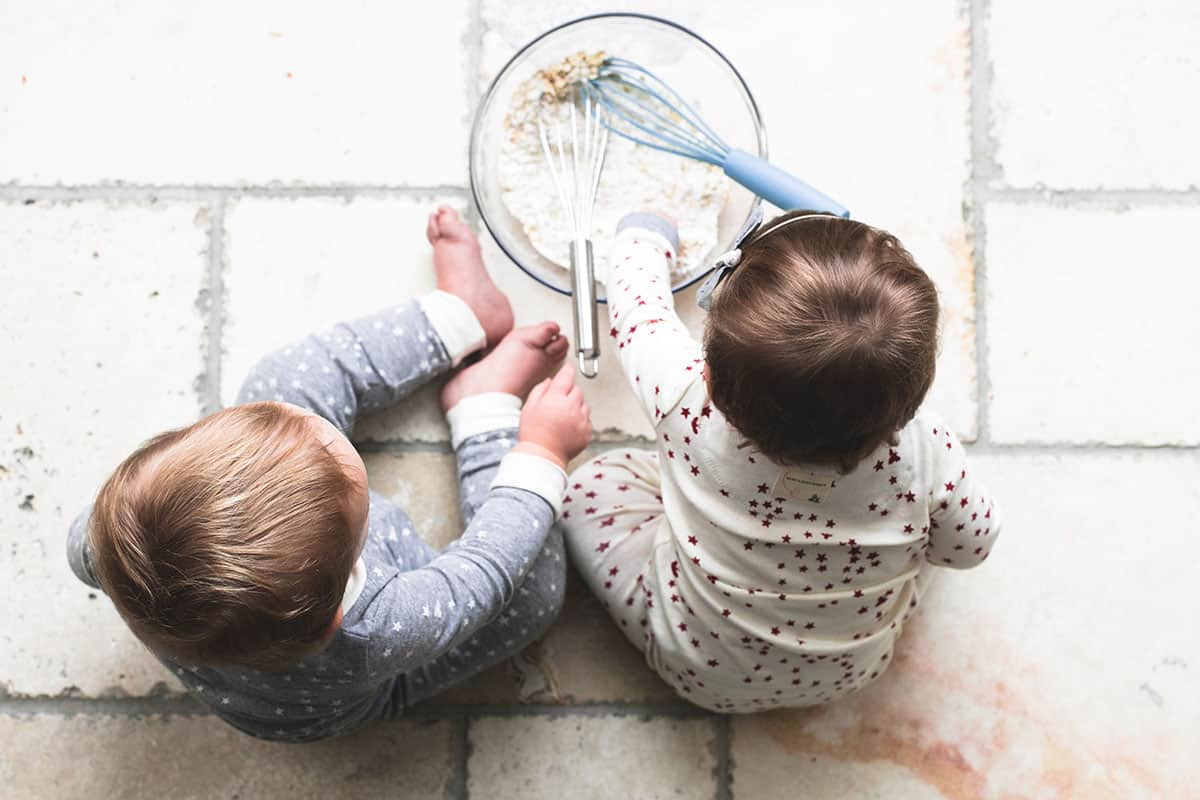 top view of two kids sitting on the floor in front of a glass bowl of pancake mix and oats with two whisks.