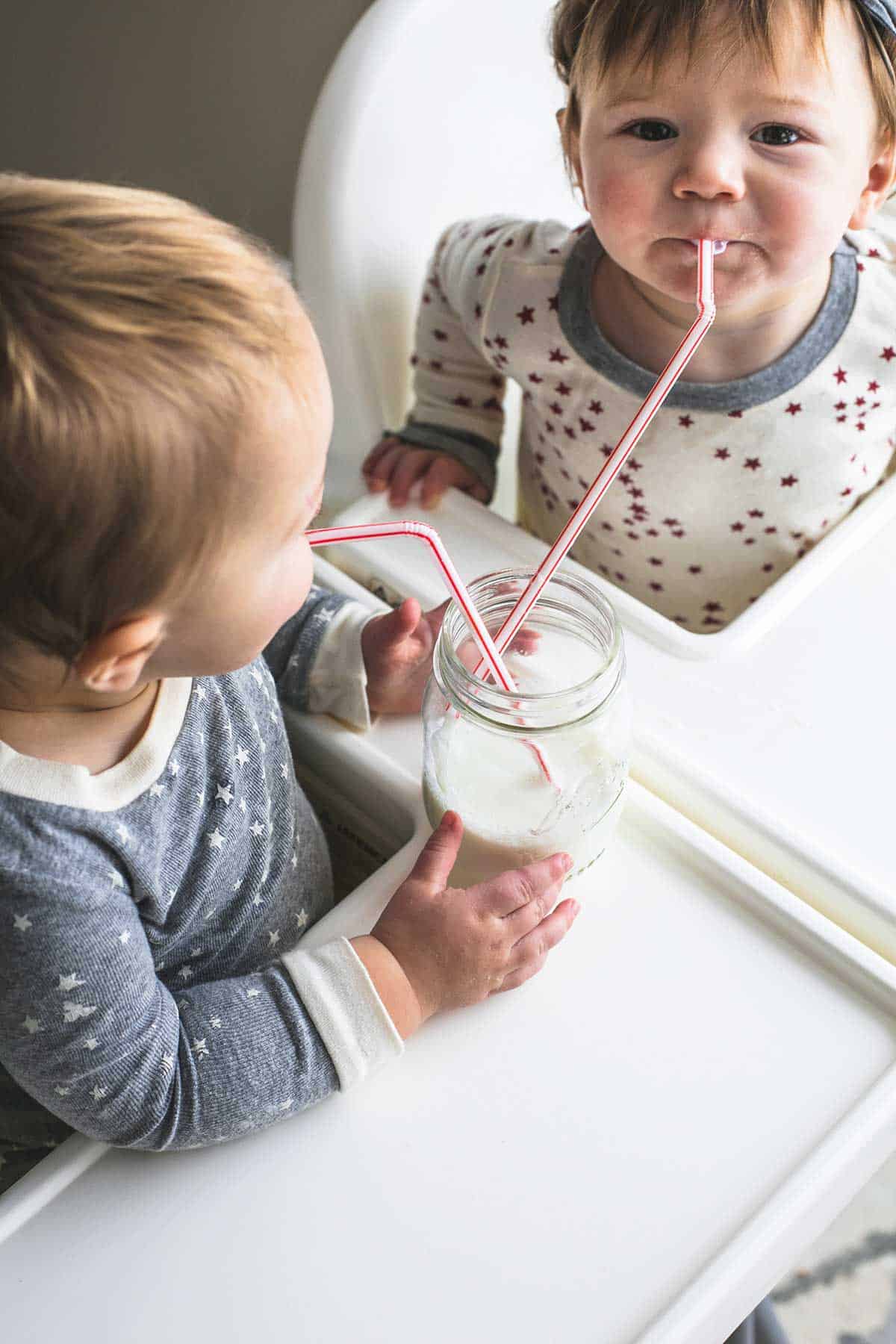 two kids sitting in high chairs both drinking out of a mason jar of milk with two straws.