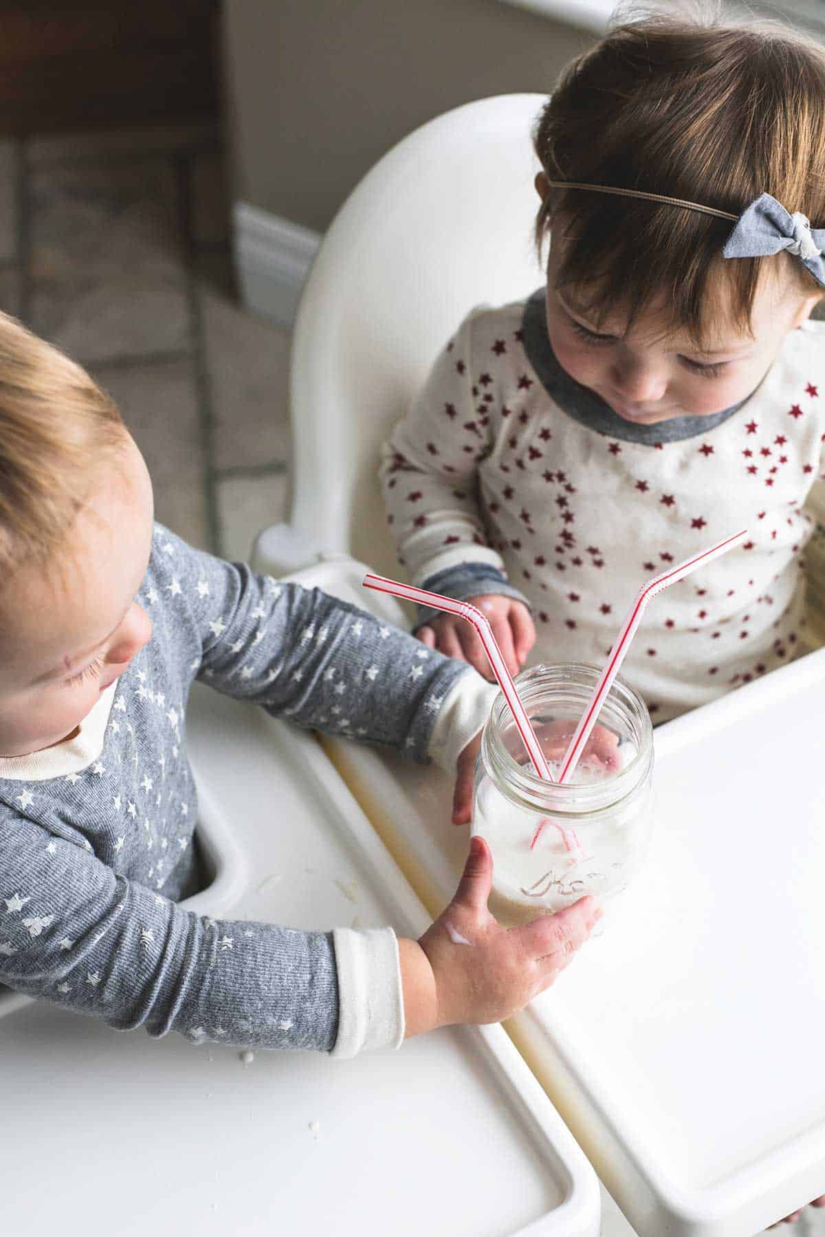 two. kids sitting in high chairs with one kid holding a mason jar of milk with two straws.