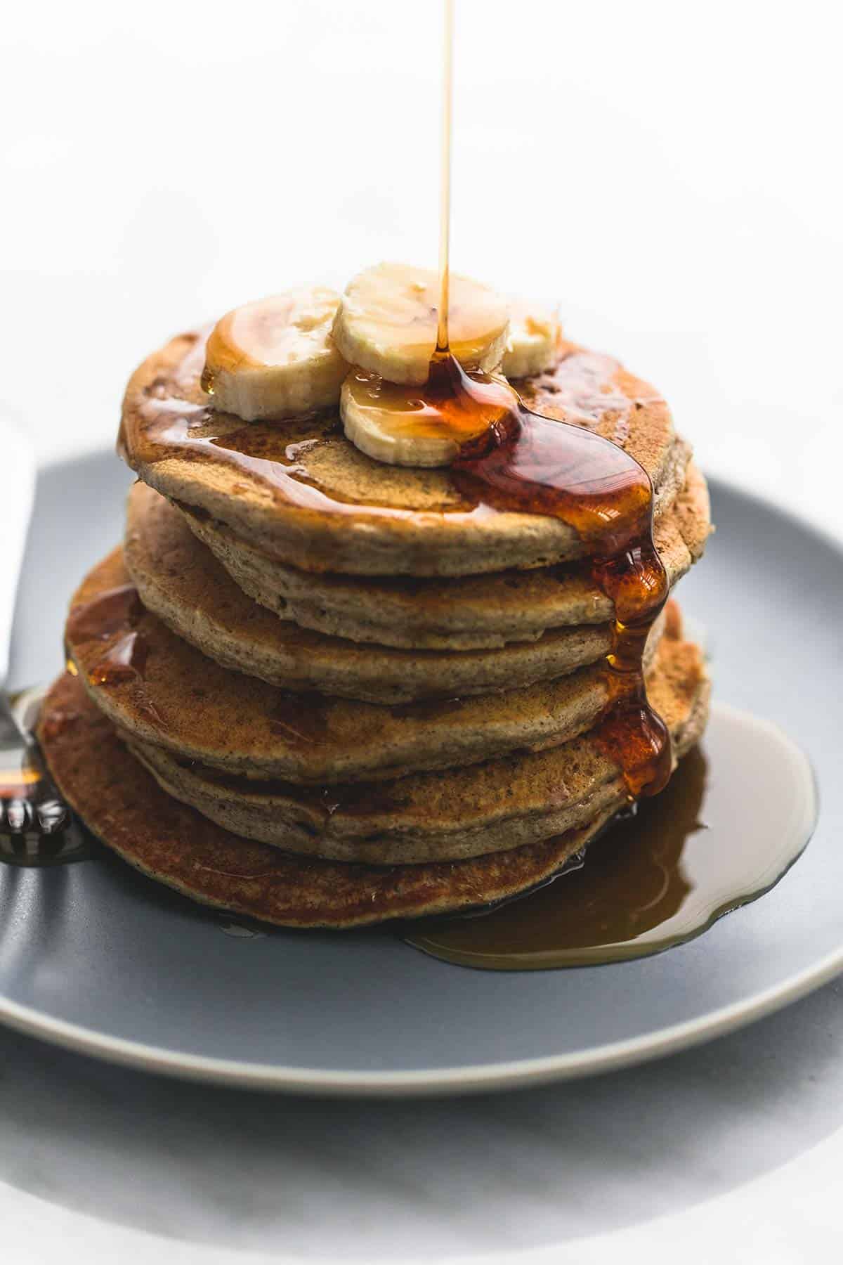 a stack of oatmeal pancakes with a fork on a plate topped with bananas with syrup being poured on top.