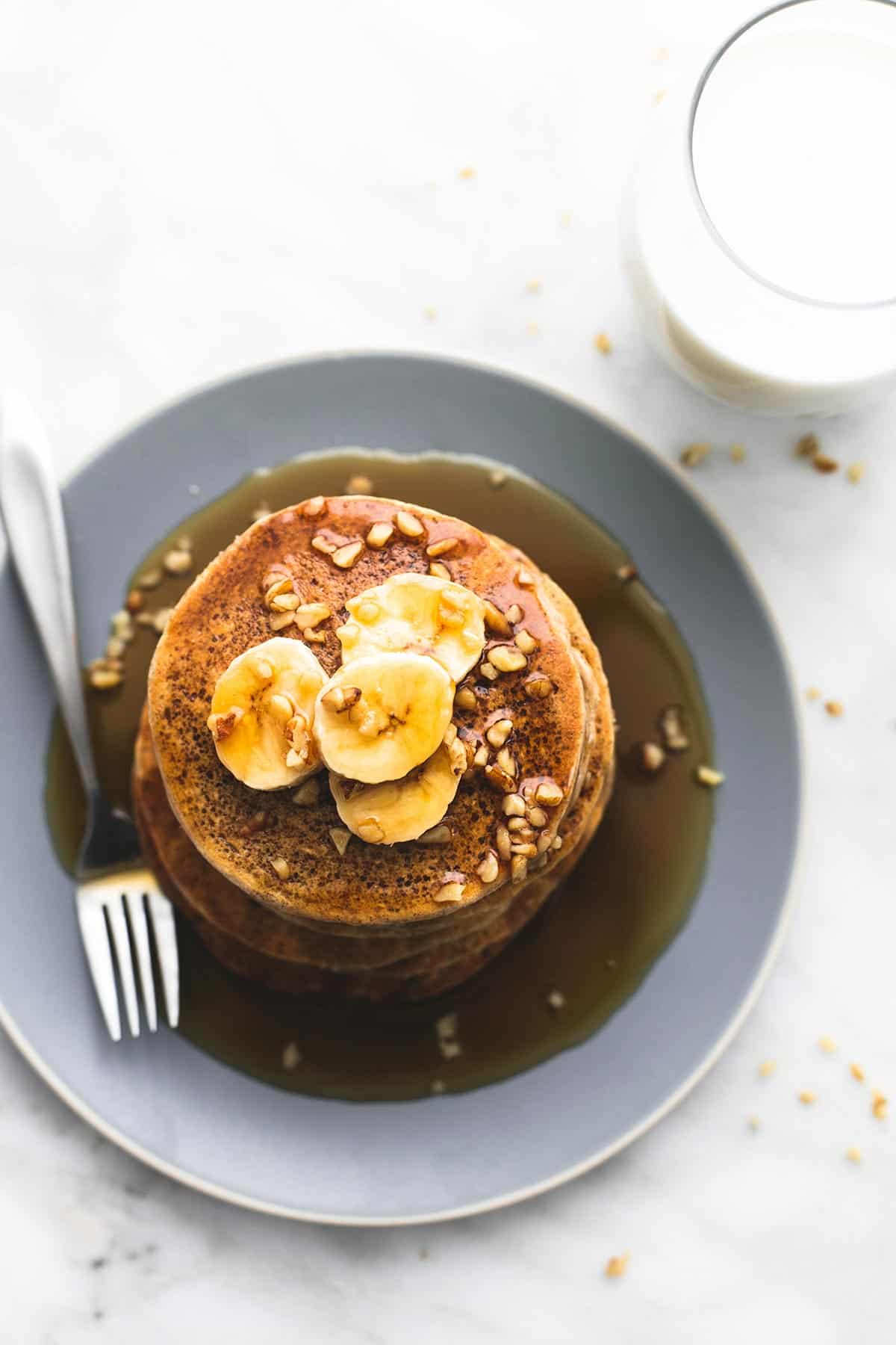 top view of a stack of oatmeal pancakes topped with bananas, chopped nuts, and syrup with a fork all on a plate with a glass of milk on the side.