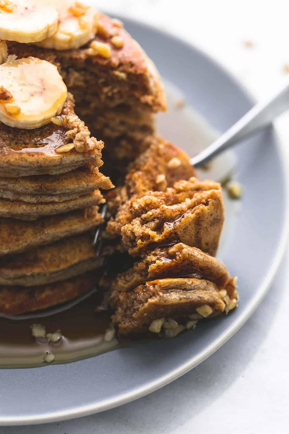 close up of a stack of oatmeal pancakes topped with bananas, chopped nuts, and syrup with a fork in between a bite and the rest of the pancakes on a plate.