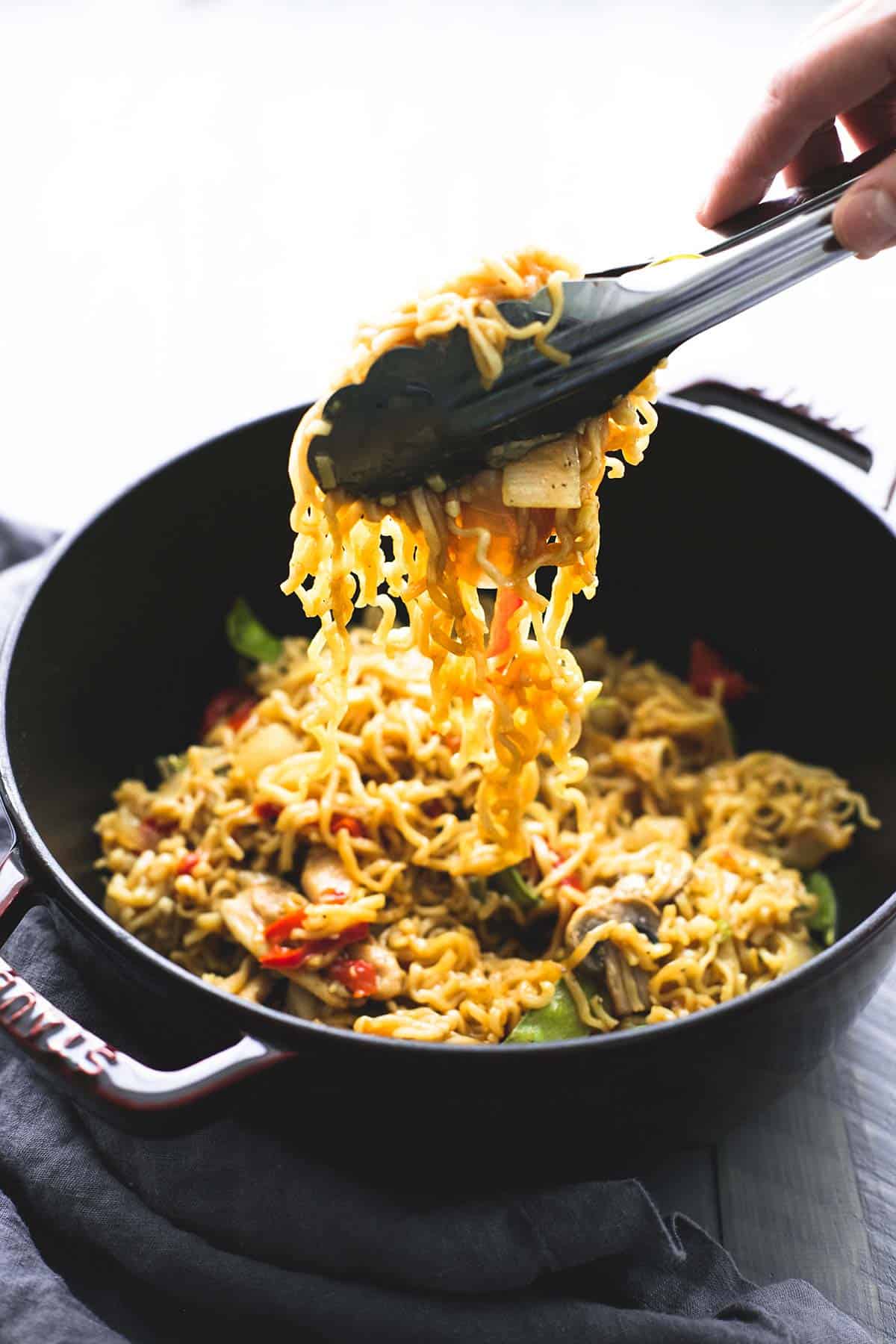 a hand picking up chicken & veggie ramen with tongs from a wok.