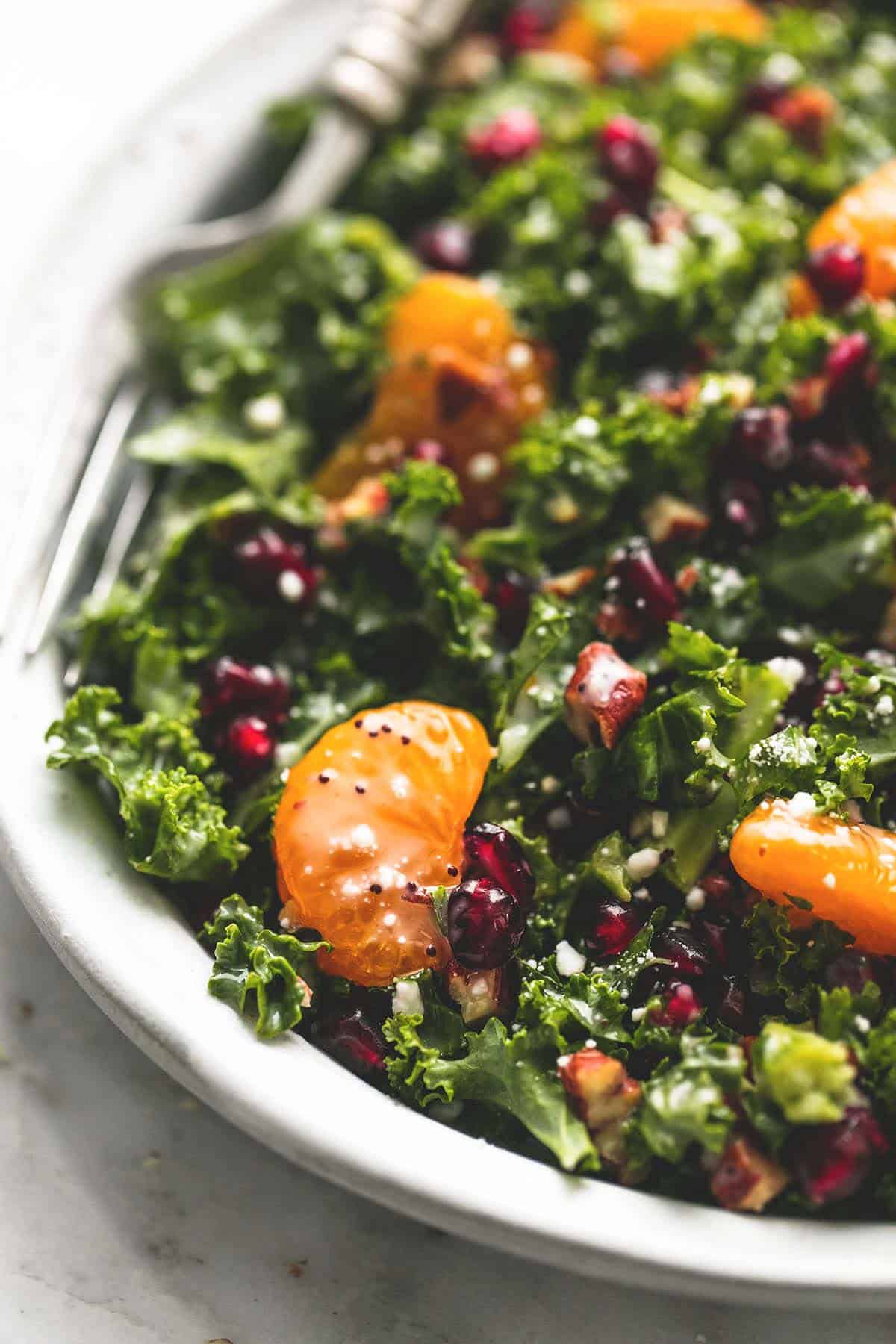 close up of mandarin pomegranate chopped kale salad with lemony poppyseed dressing with a fork on a plate.
