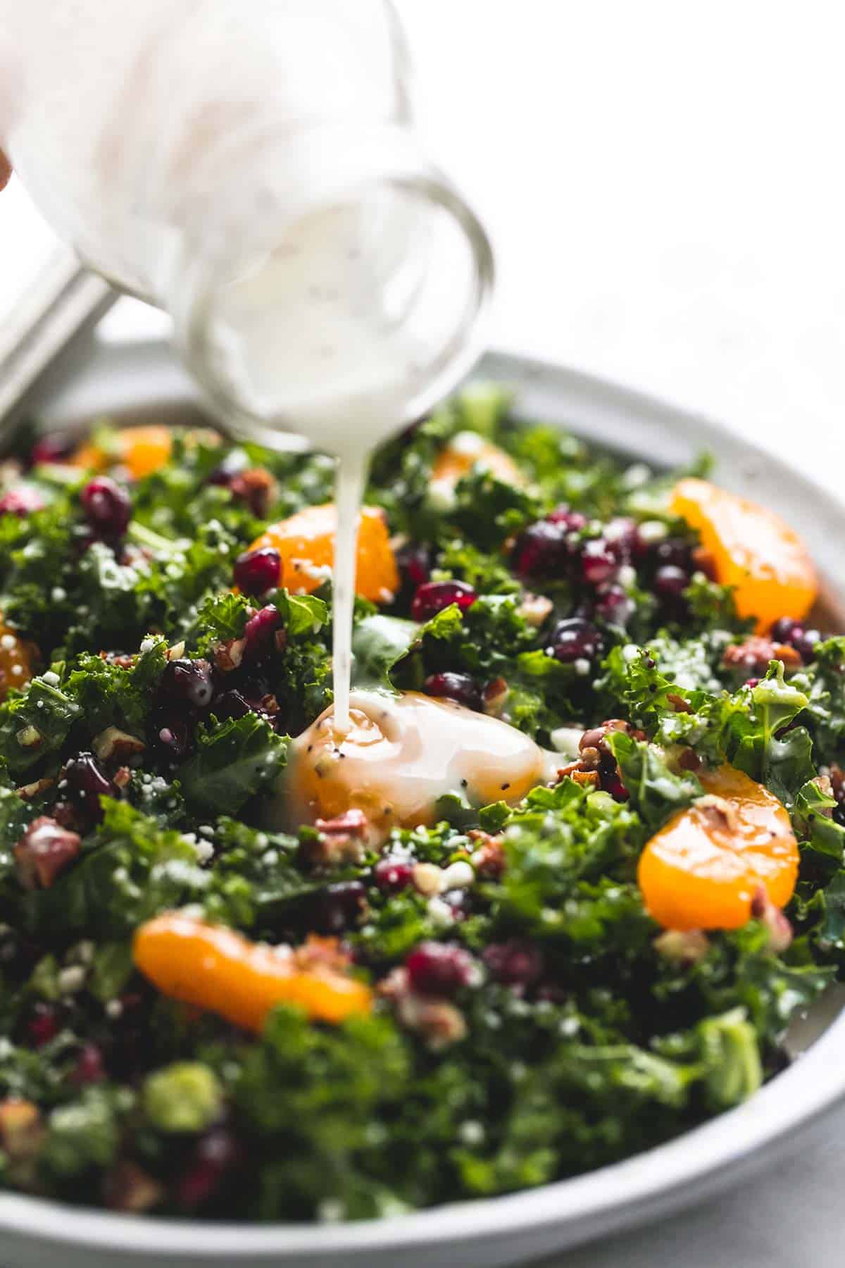 close up of lemony poppyseed dressing being poured on top of mandarin pomegranate chopped kale salad on a plate.