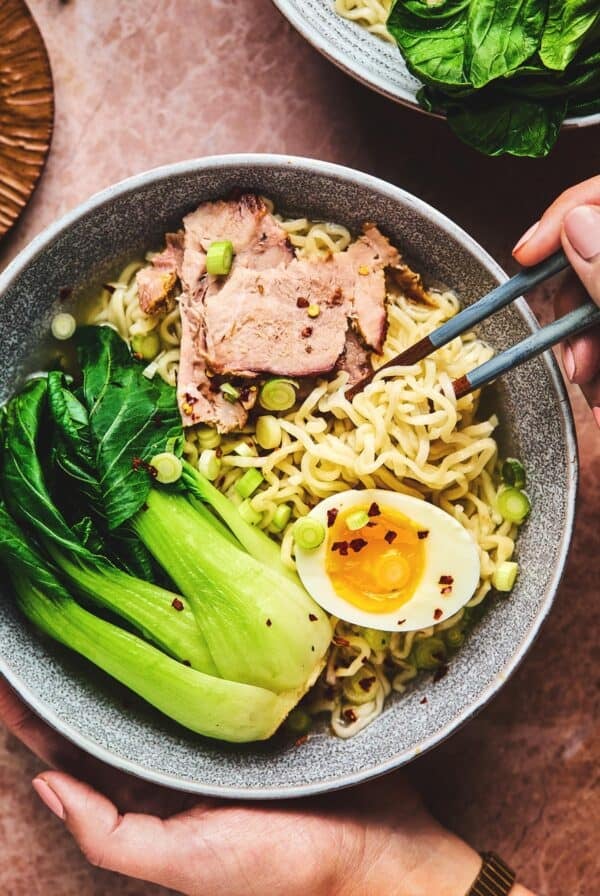 Hand holding bowl of pork ramen noodles and chopsticks.