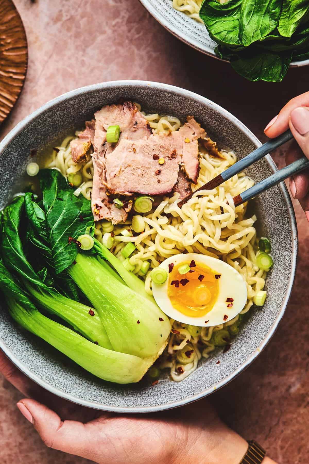 Hand holding bowl of pork ramen noodles and chopsticks.