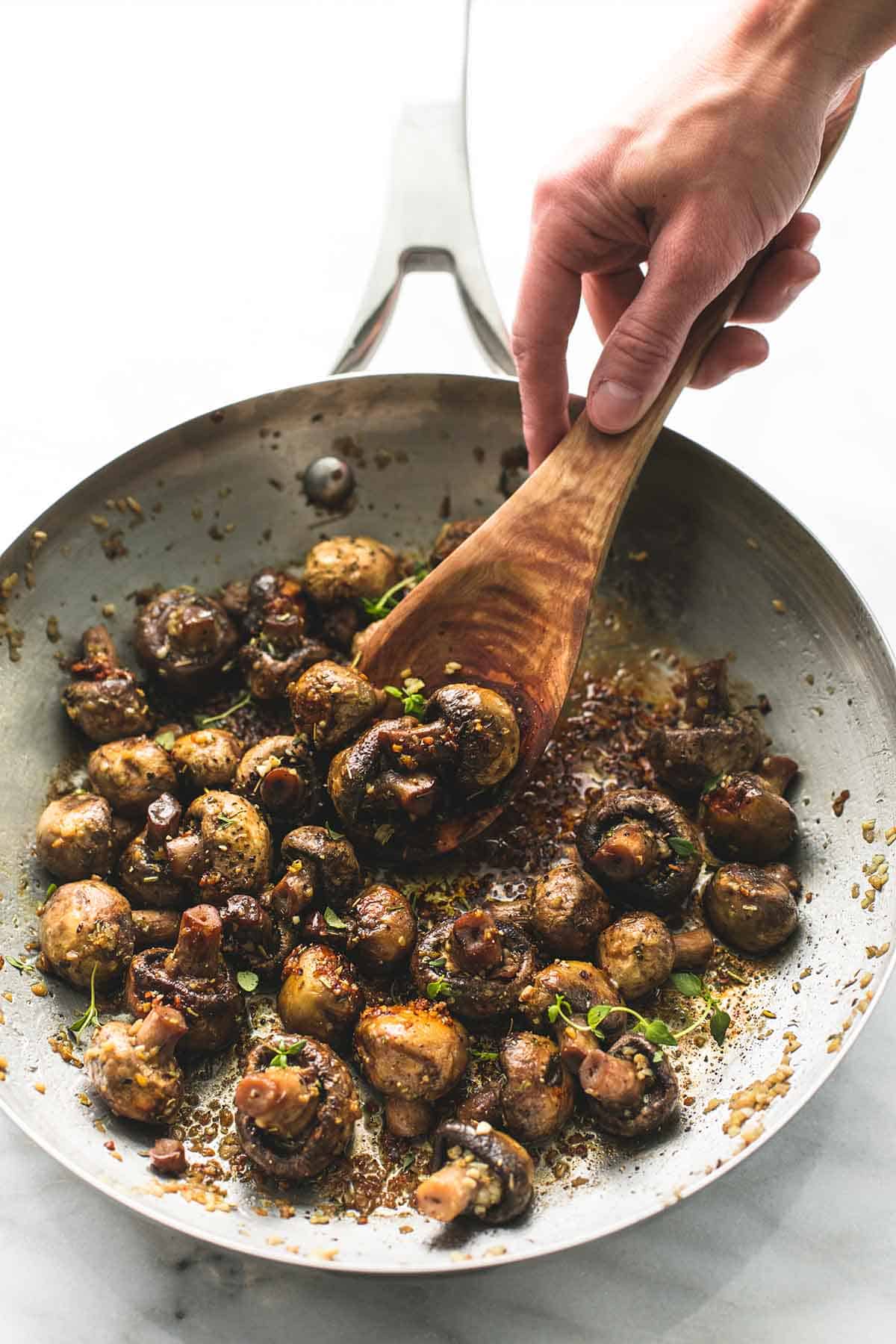 a hand grabbing some sautéed garlic butter mushrooms with a wooden serving spoon from a skillet.