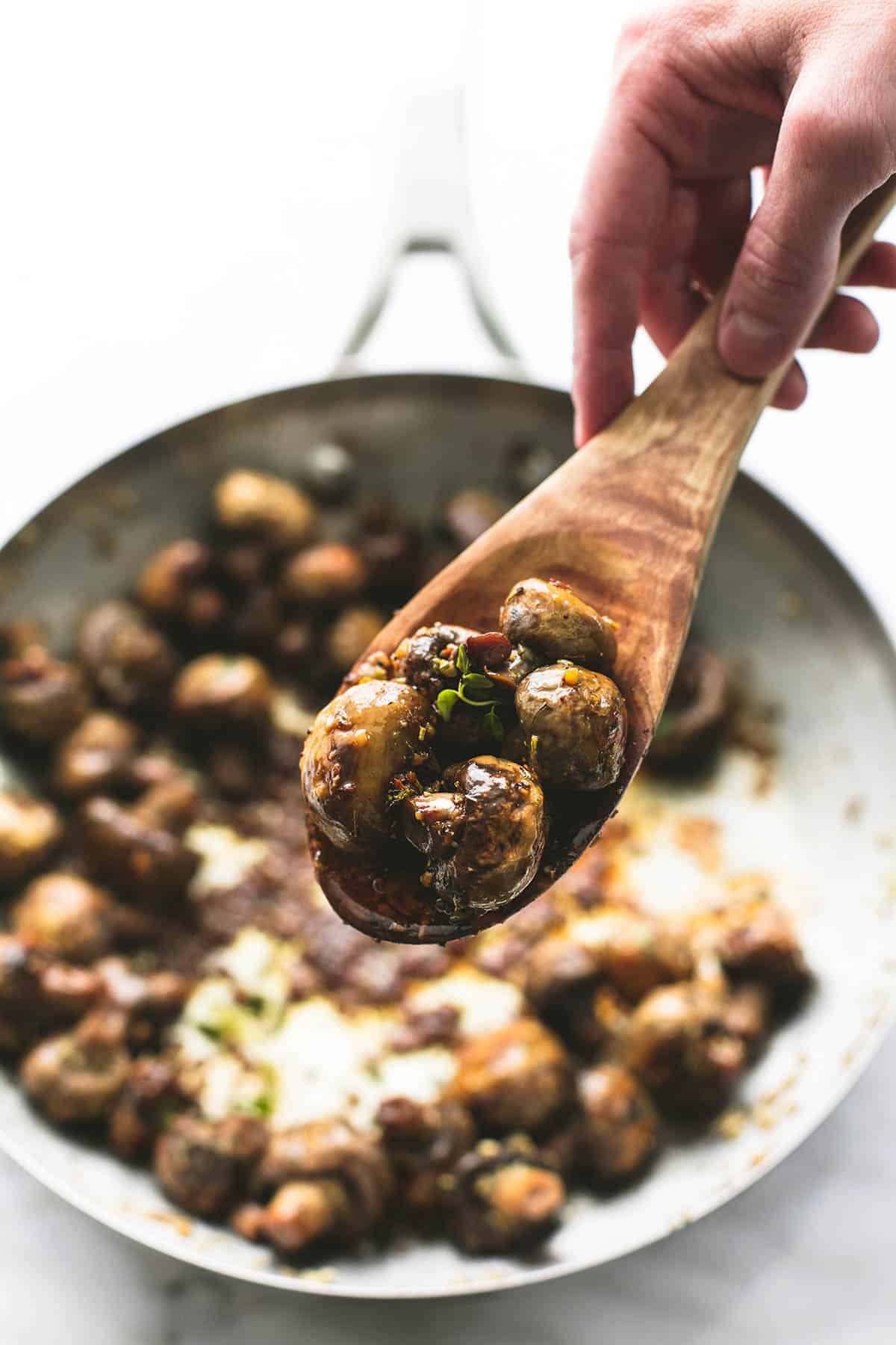 a hand holding a wooden serving spoon with garlic butter mushrooms on it above a skillet of more mushrooms.