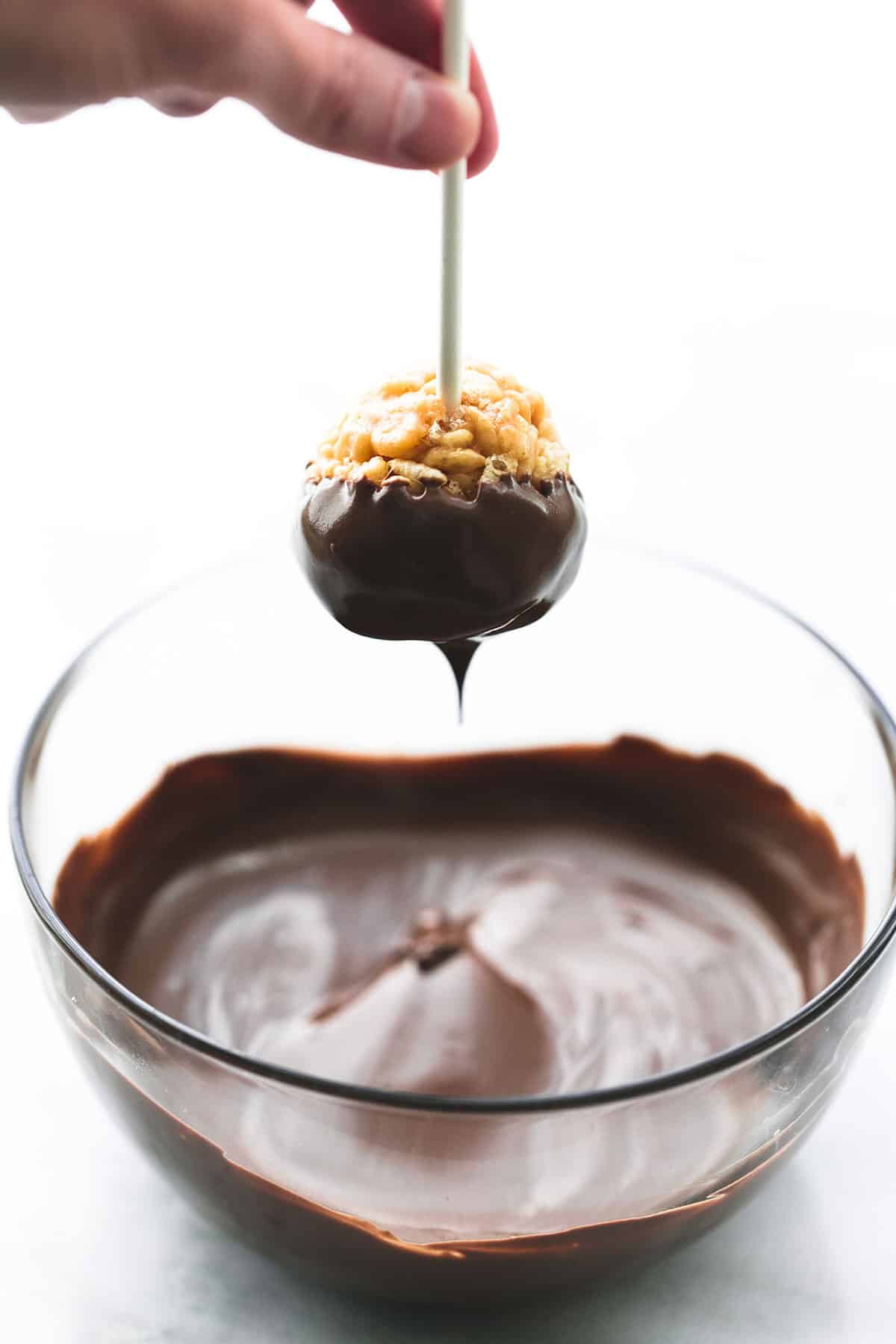 a hand holding a scotcheroo on a stick just dipped in chocolate above a bowl of chocolate.