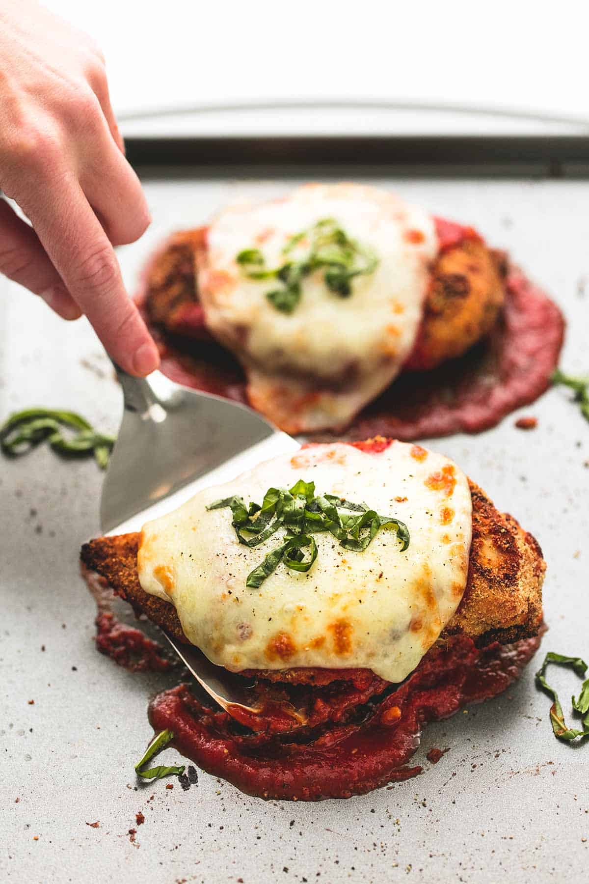 a hand picking up a piece of baked chicken parmesan from a baking sheet with a spatula.