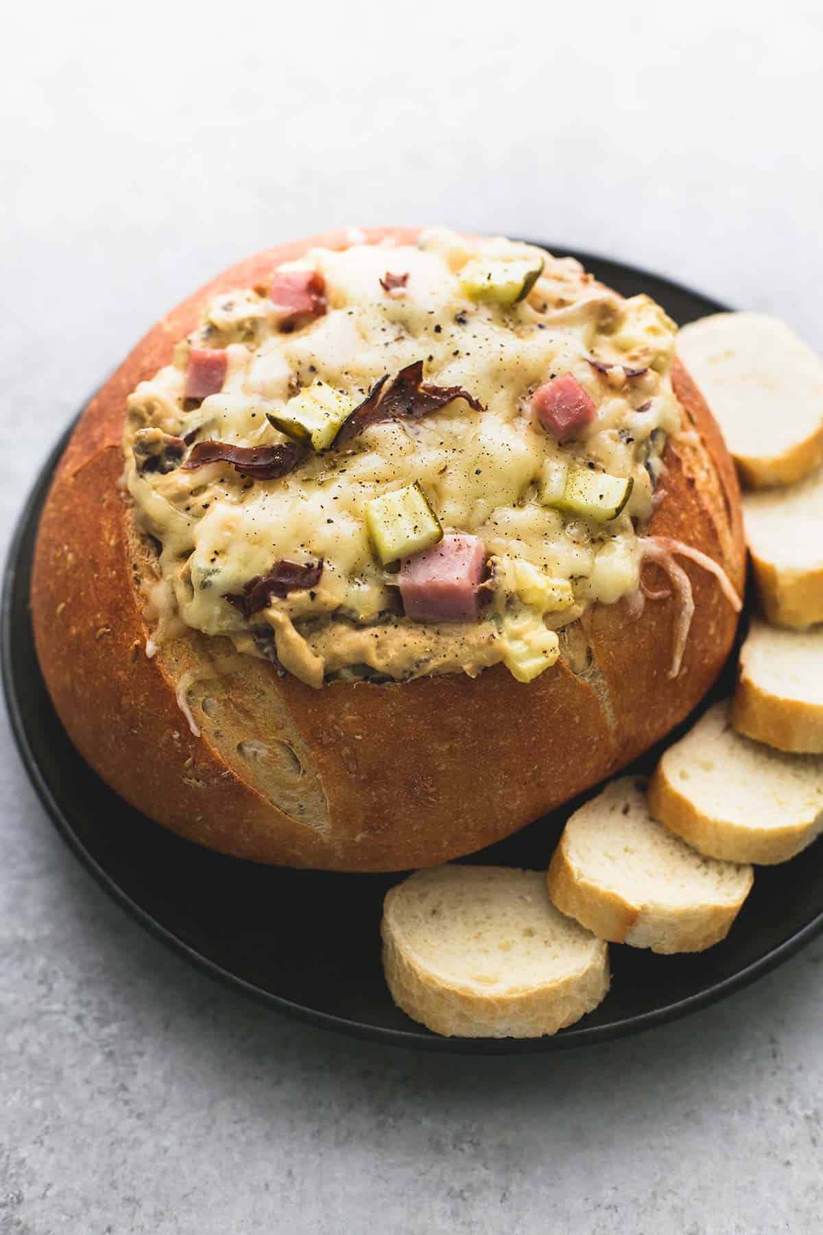 a bread bowl of Miami Cuban dip with slices of Italian baguettes on the side all on a plate.