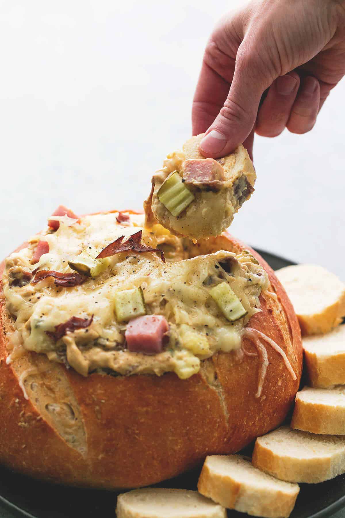a hand holding a slice of Italian baguette just dipped in Miami Cuban dip above a bread bowl of dip with more slices of bread on the side all on a plate.