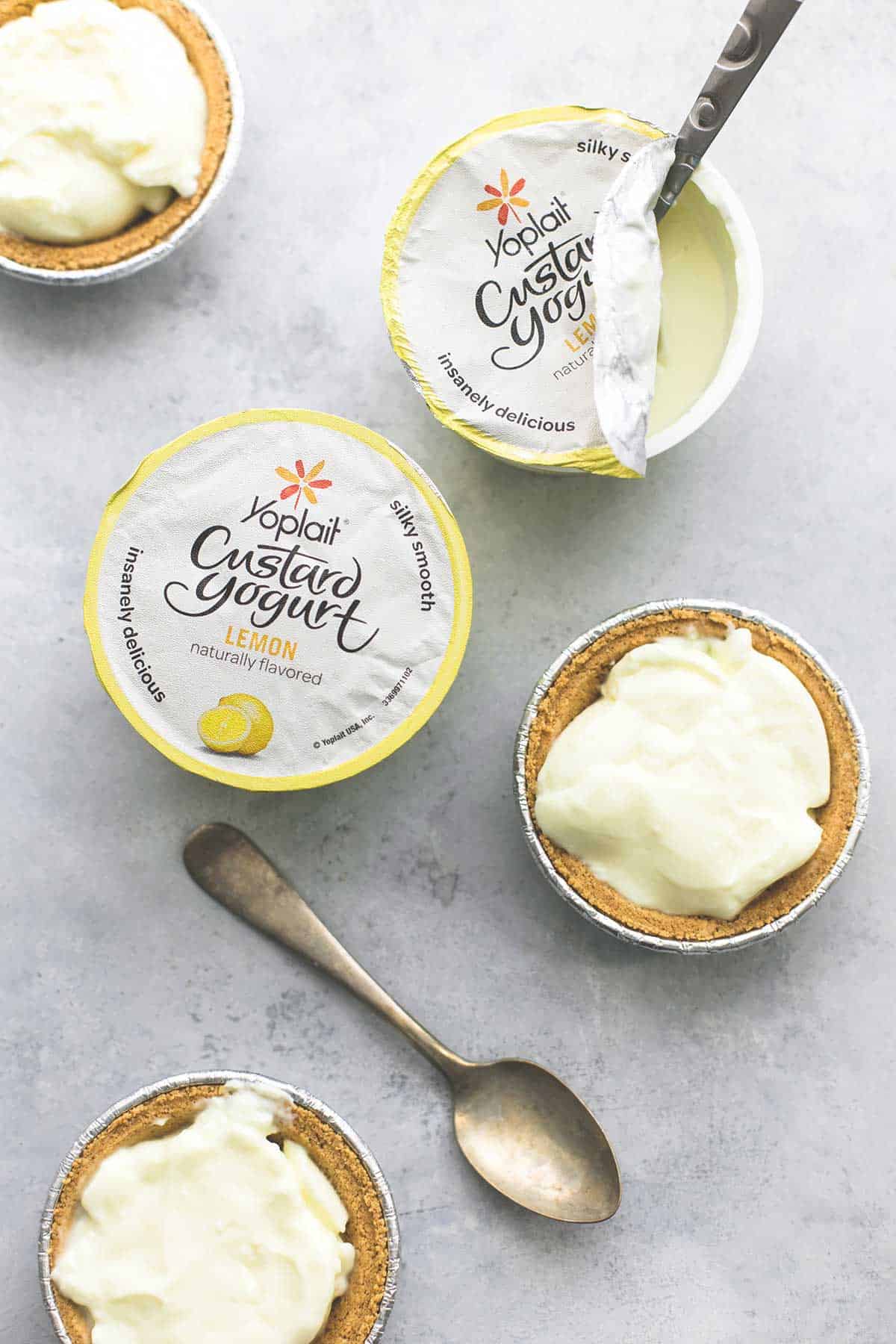 top view of lemon custard yogurt mini pies and yogurt container with a spoon on the side of one and the other opened with a spoon in it.