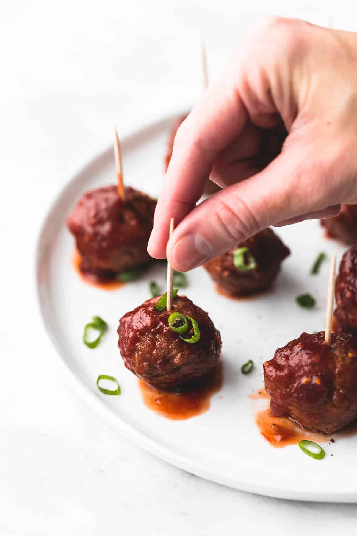 a hand picking up a slow cooker party meatball from a plate of meatballs with toothpicks in them.