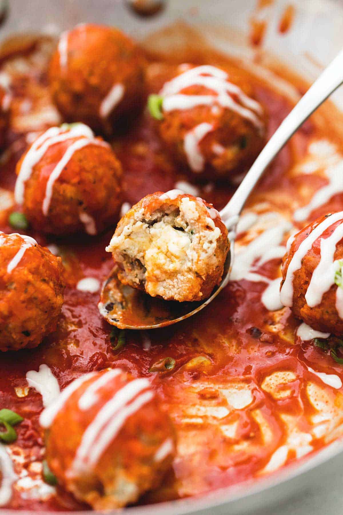 close up of a blue cheese stuffed buffalo chicken meatball with a bite missing on a spoon with more meatballs around it drizzled with blue cheese dressing on top in a pan.