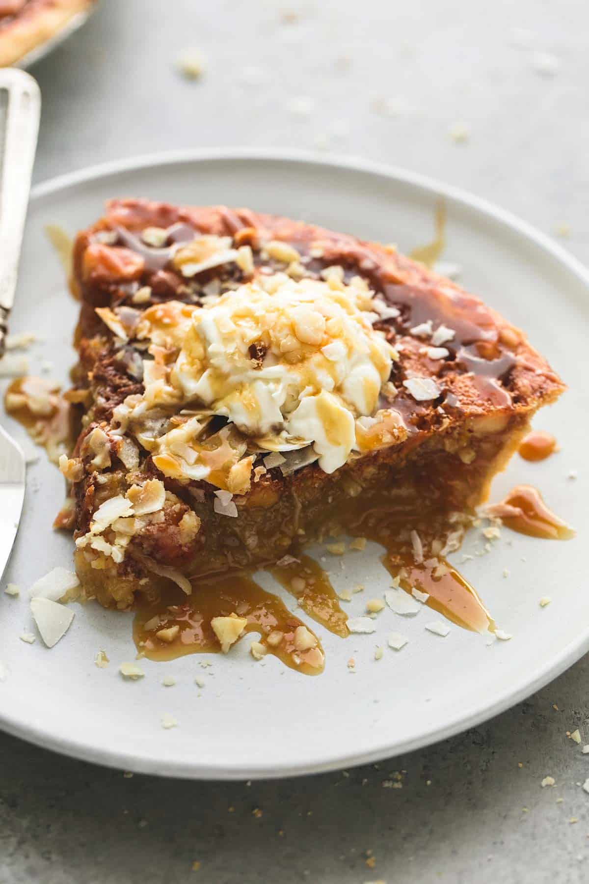 a slice of caramel coconut macadamia nut pie topped with whipped cream coconut shavings and caramel sauce on a plate.
