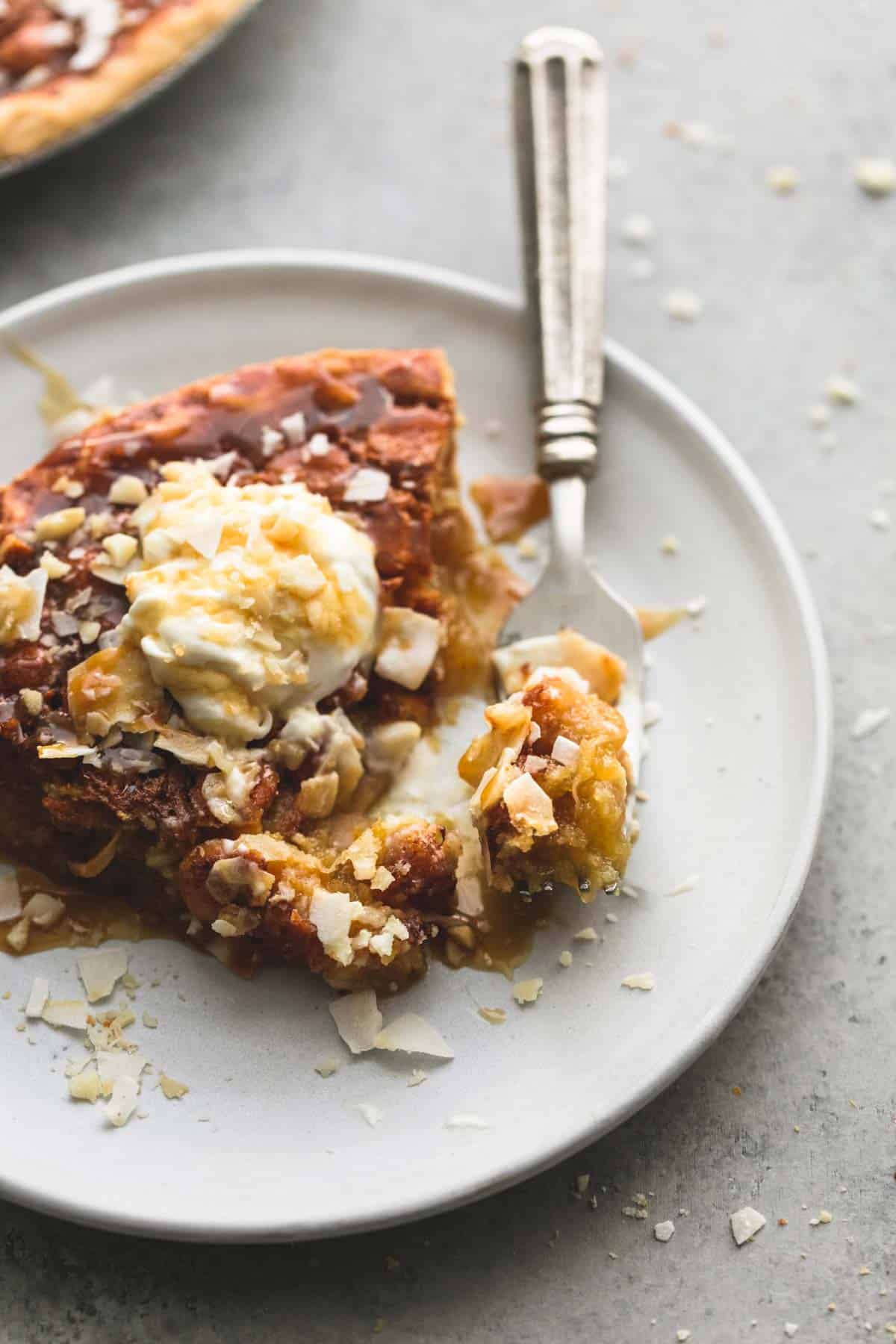 a slice of caramel coconut macadamia nut pie topped with whipped cream, coconut shavings and caramel sauce with a bite on a fork on a plate.