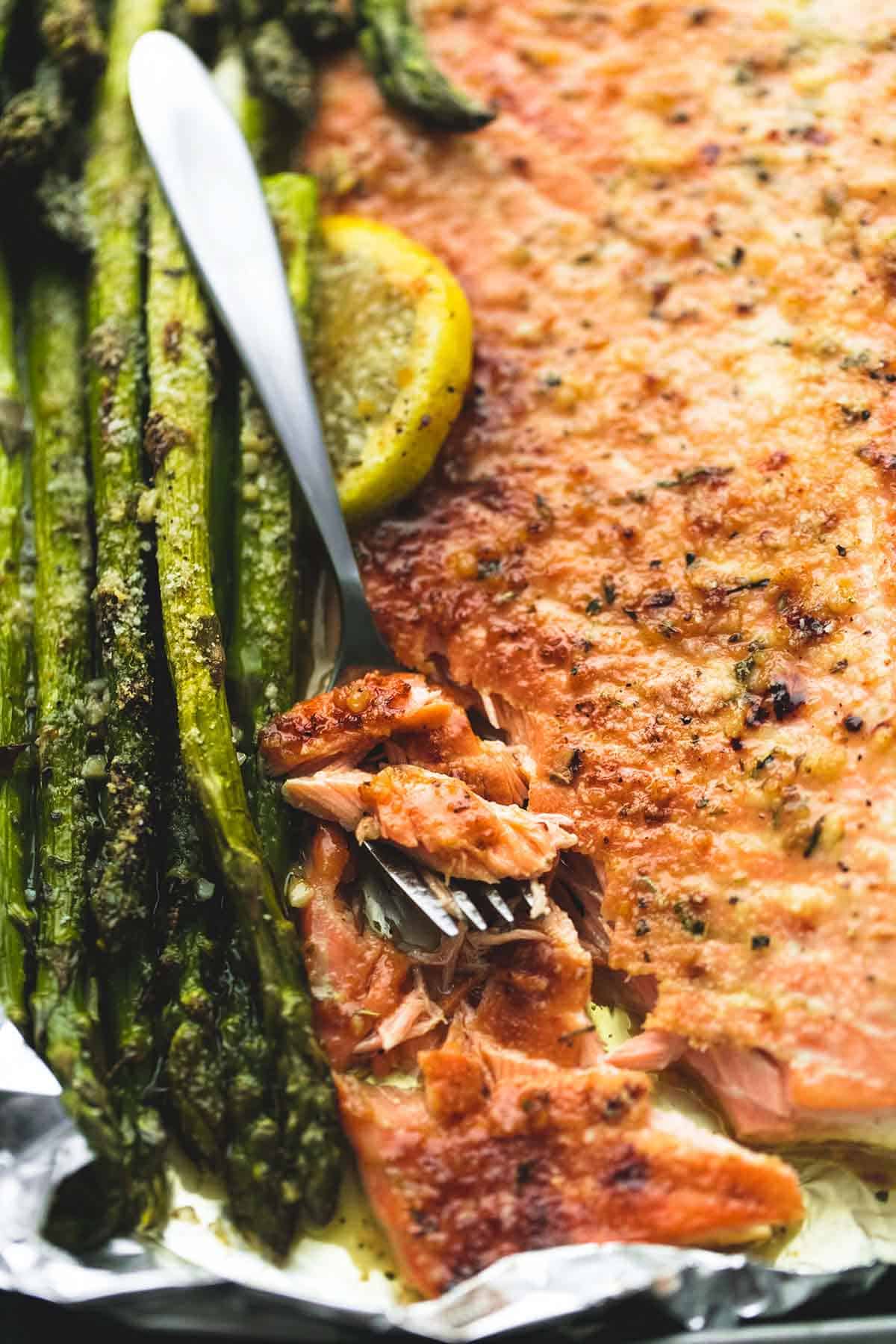 close up top view of baked lemon parmesan salmon & asparagus in foil with a bite on a fork sitting on top of the salmon.