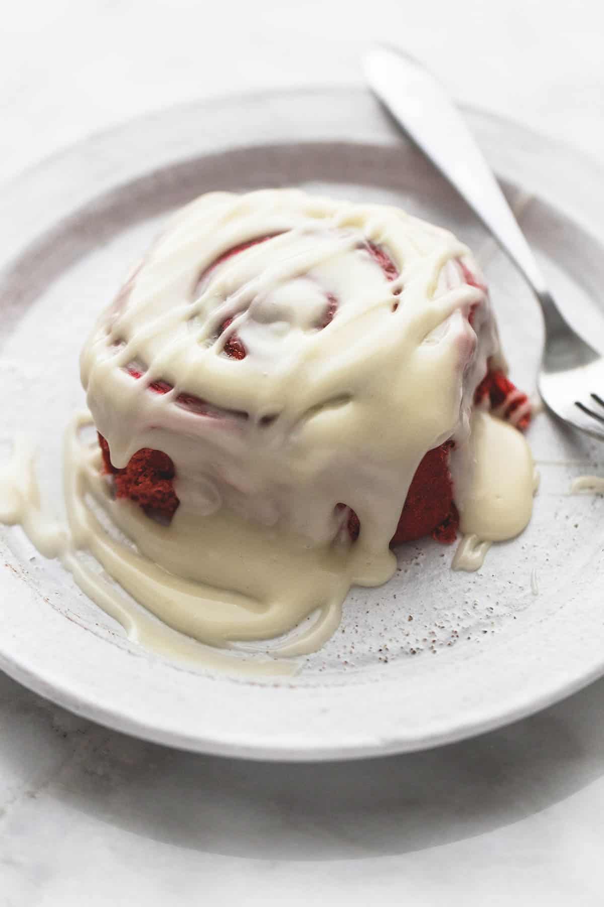 a red velvet cinnamon roll with brown butter cream cheese frosting with a fork on a plate.