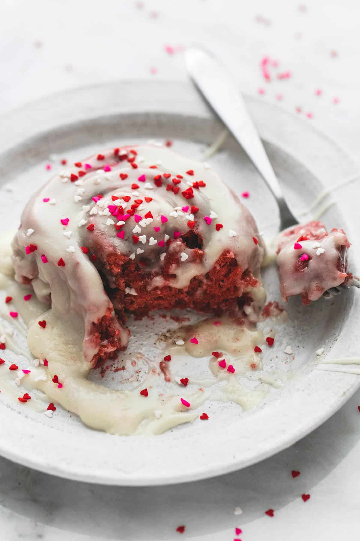 a red velvet cinnamon roll with brown butter cream cheese frosting with a bite on a fork topped with sprinkles on a plate.