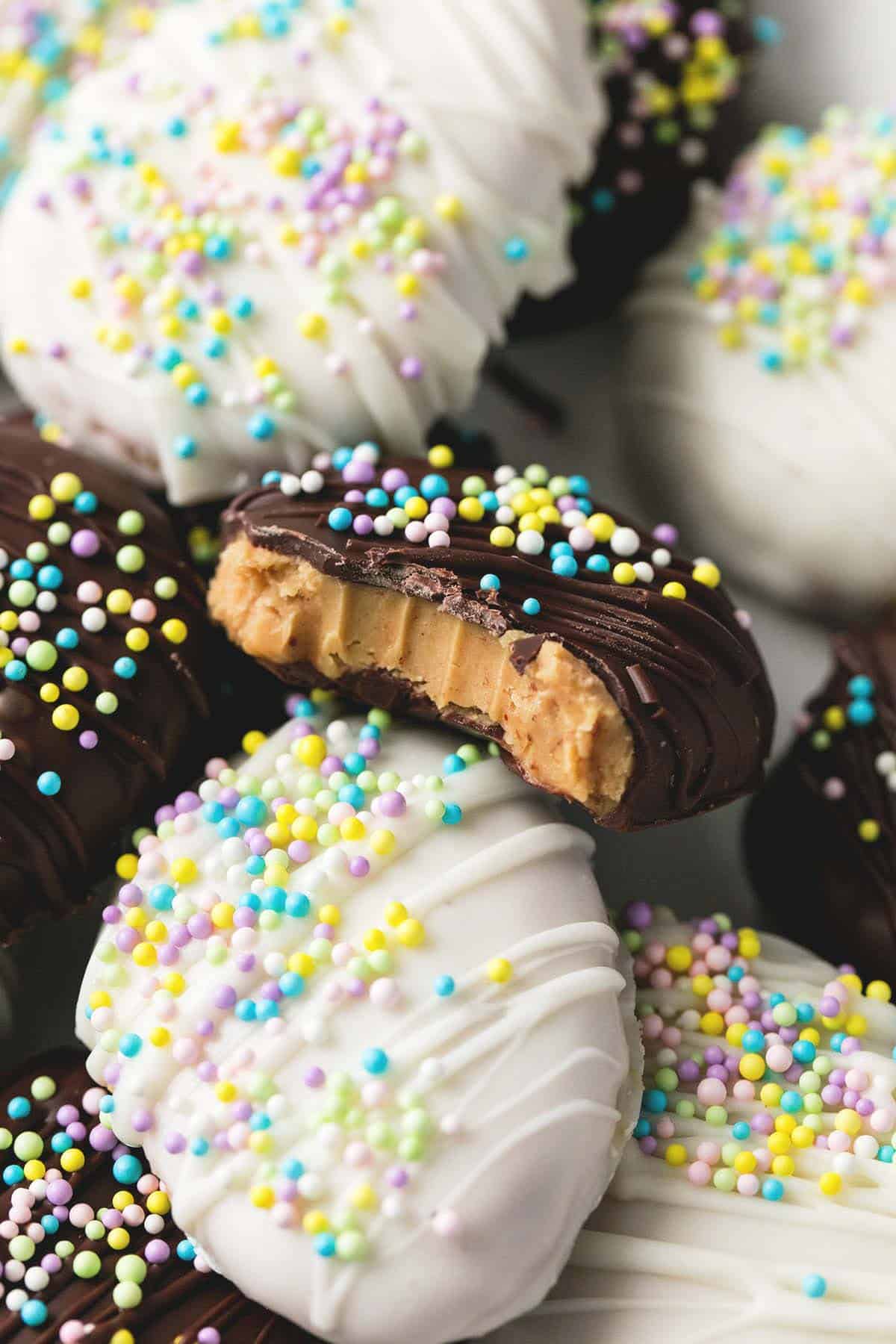 close up of a pile of copycat Reese's creamy peanut butter eggs both white and milk chocolate with sprinkles on top with the top milk chocolate egg missing a bite.