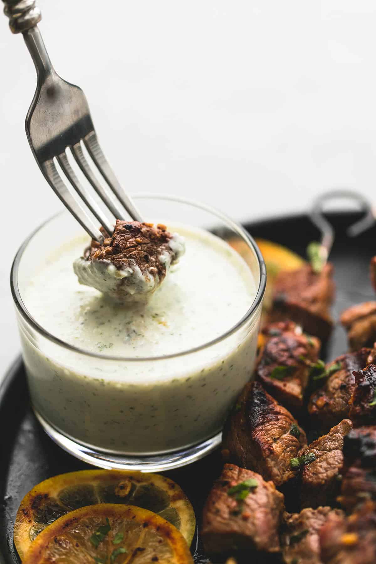 a fork with a piece of beef being dipped in tzatziki sauce with beef Souvlaki kebabs on the side.