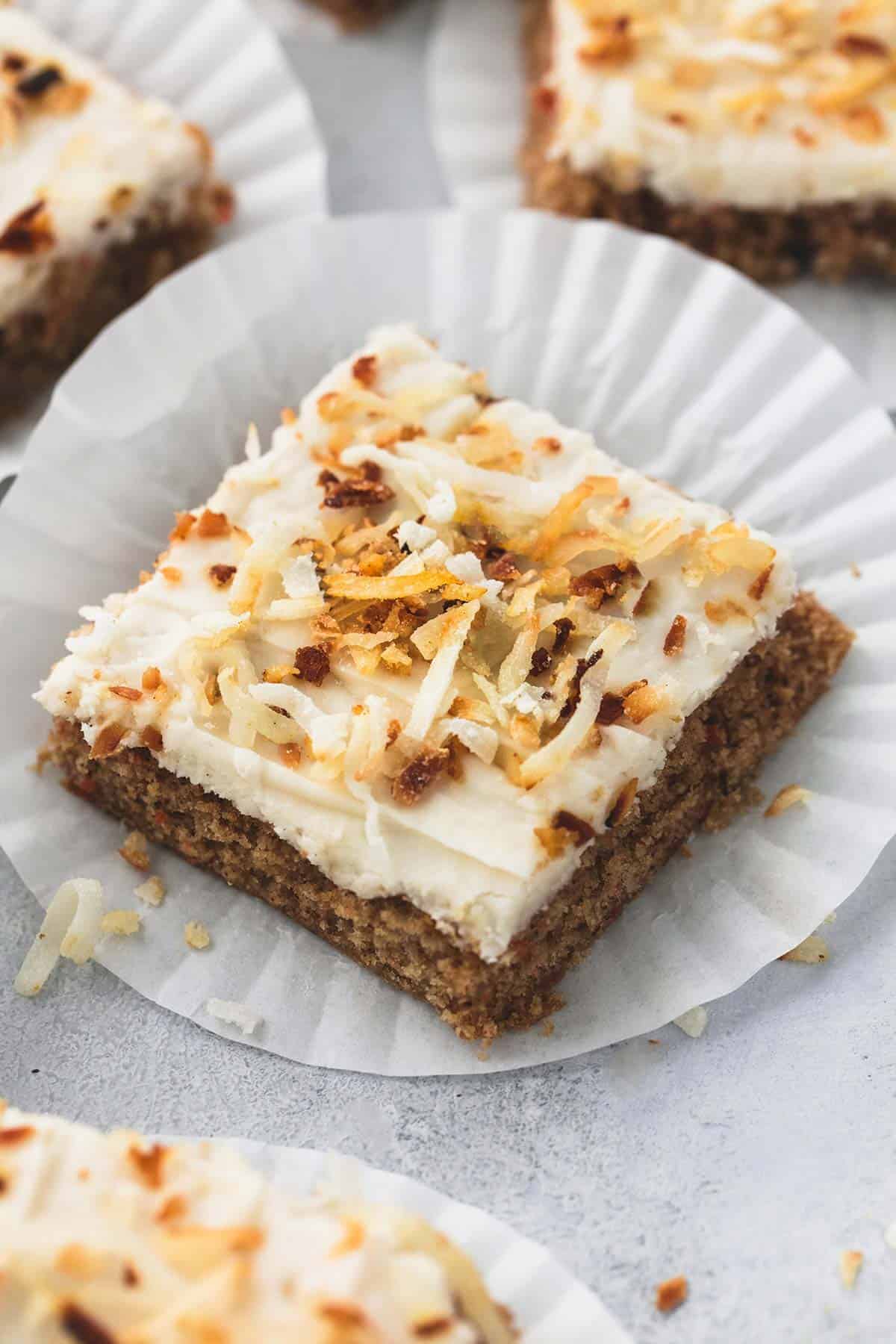 The BEST super moist carrot cake bars recipe (starts with a cake mix!) topped with coconut cream cheese frosting. | lecremedelacrumb.com a carrot cake bar with coconut cream cheese frosting on a cupcake liner with more bars on cupcake liners around it.