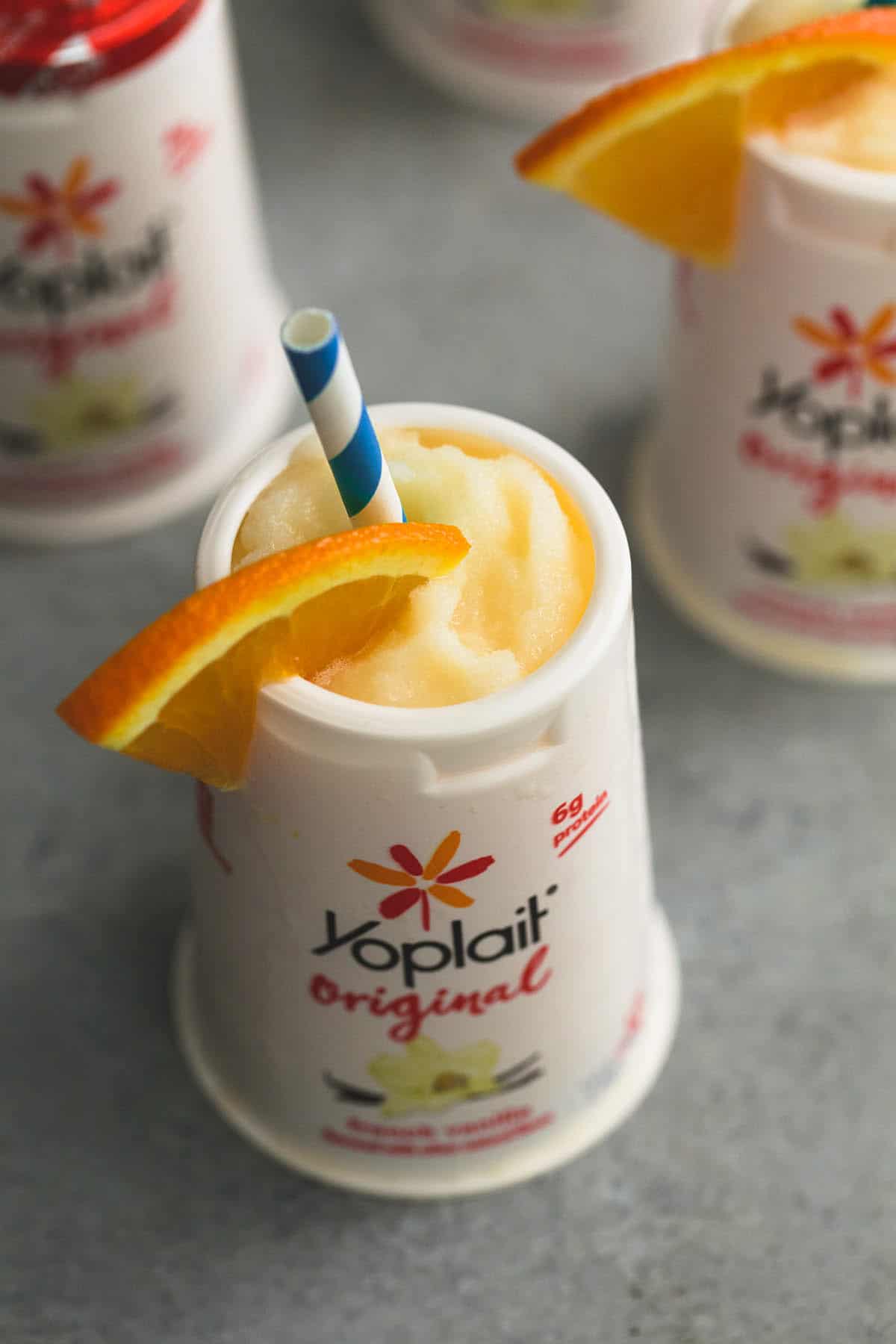 orange julius in a Yoplait container with a orange slice and straw with more Yoplait containers in the background.