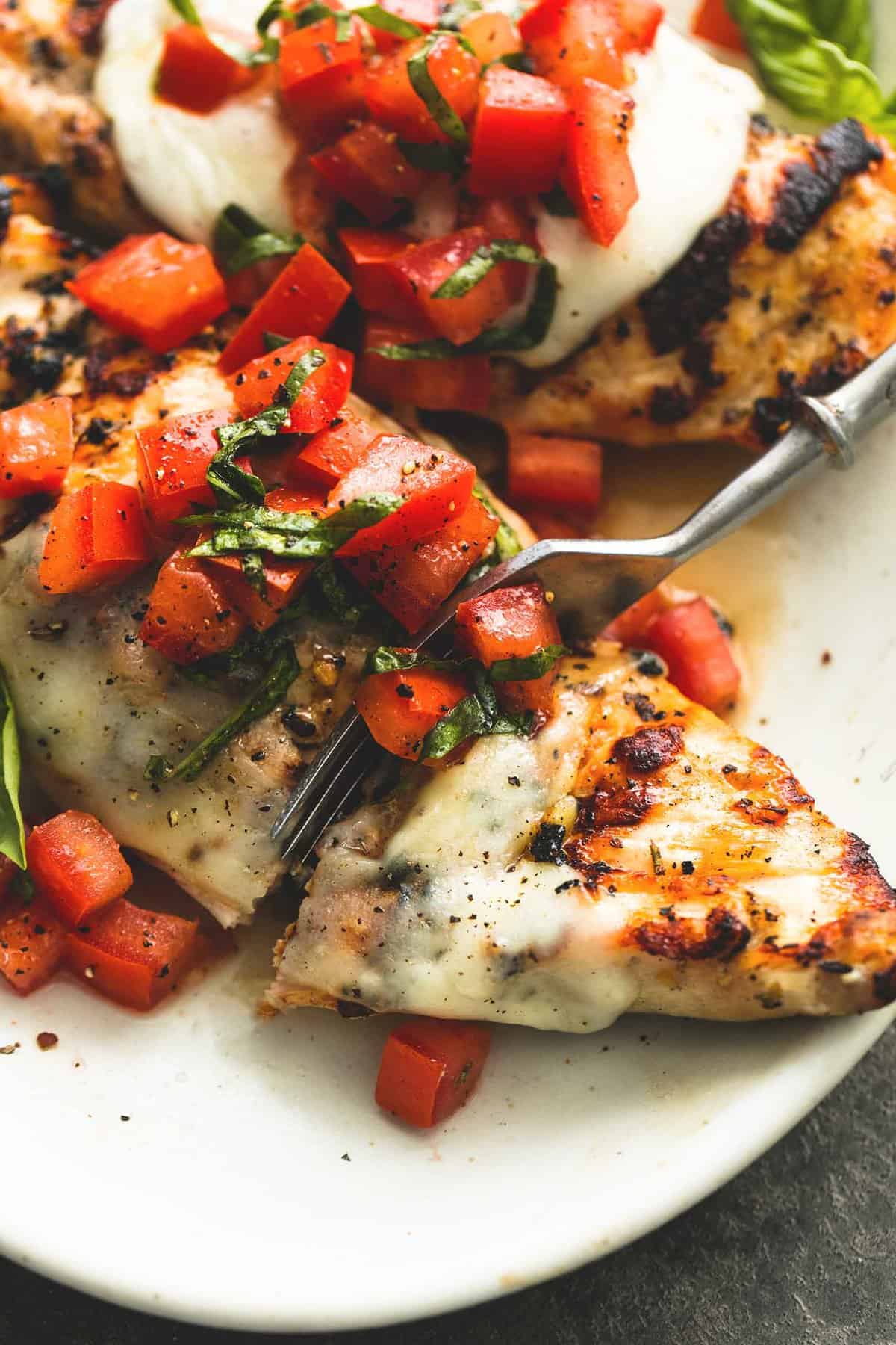 close up of grilled bruschetta chicken on a plate with a fork cutting into a piece of chicken.