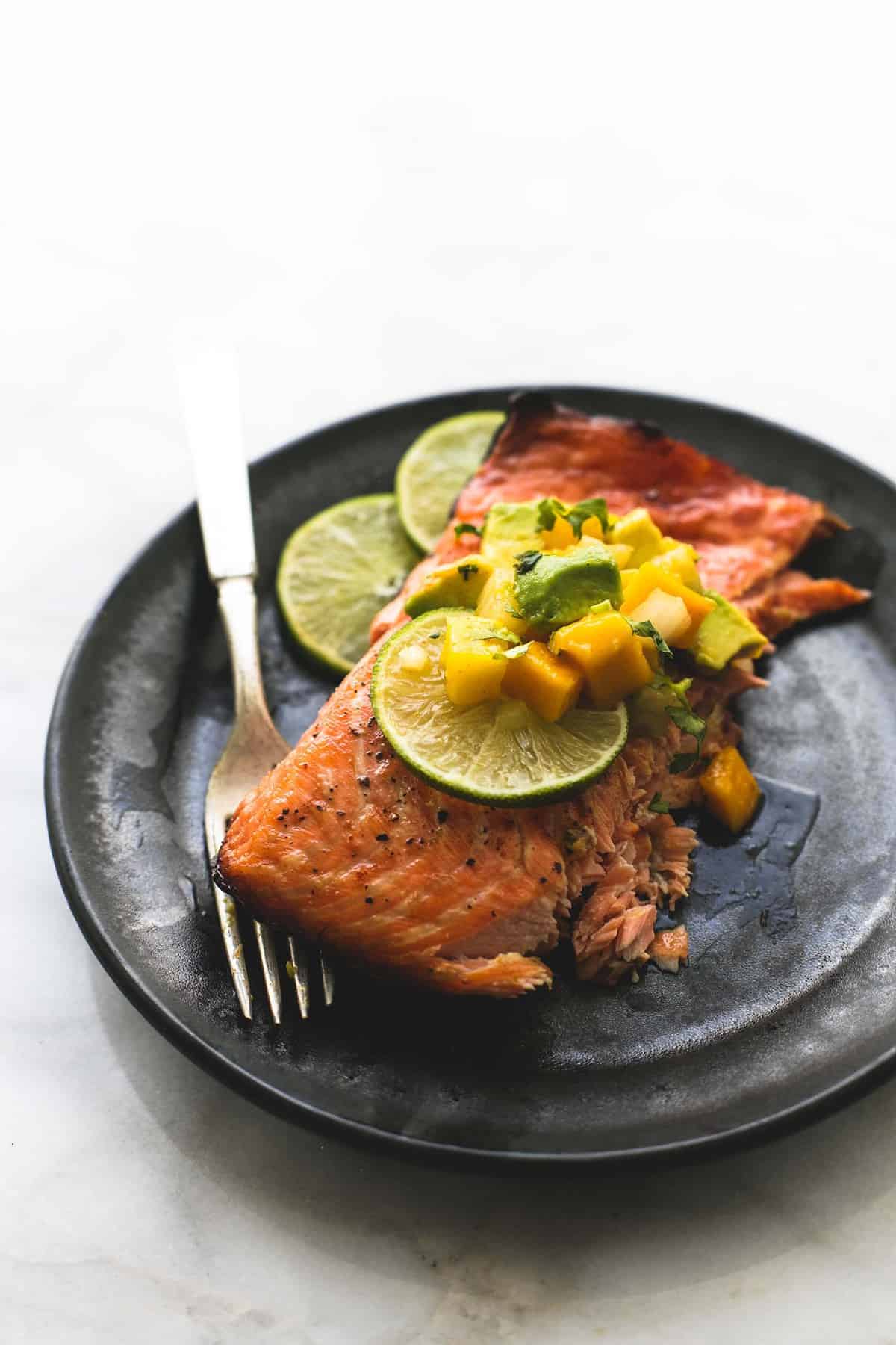 a piece of grilled honey lime salmon with mango avocado salsa and lime slices with a fork on a plate.