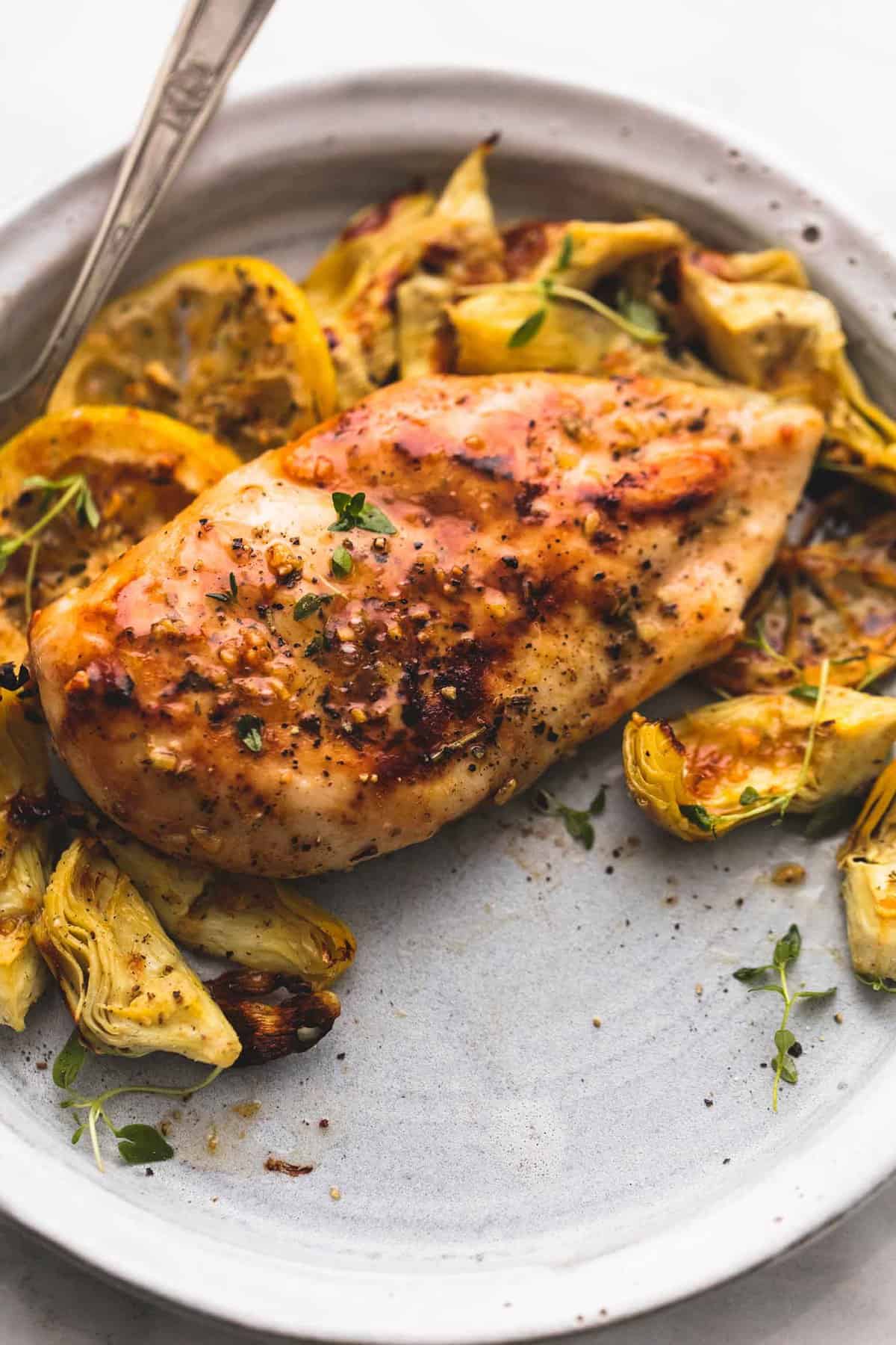 close up of baked lemon chicken and artichokes with a fork on a plate.