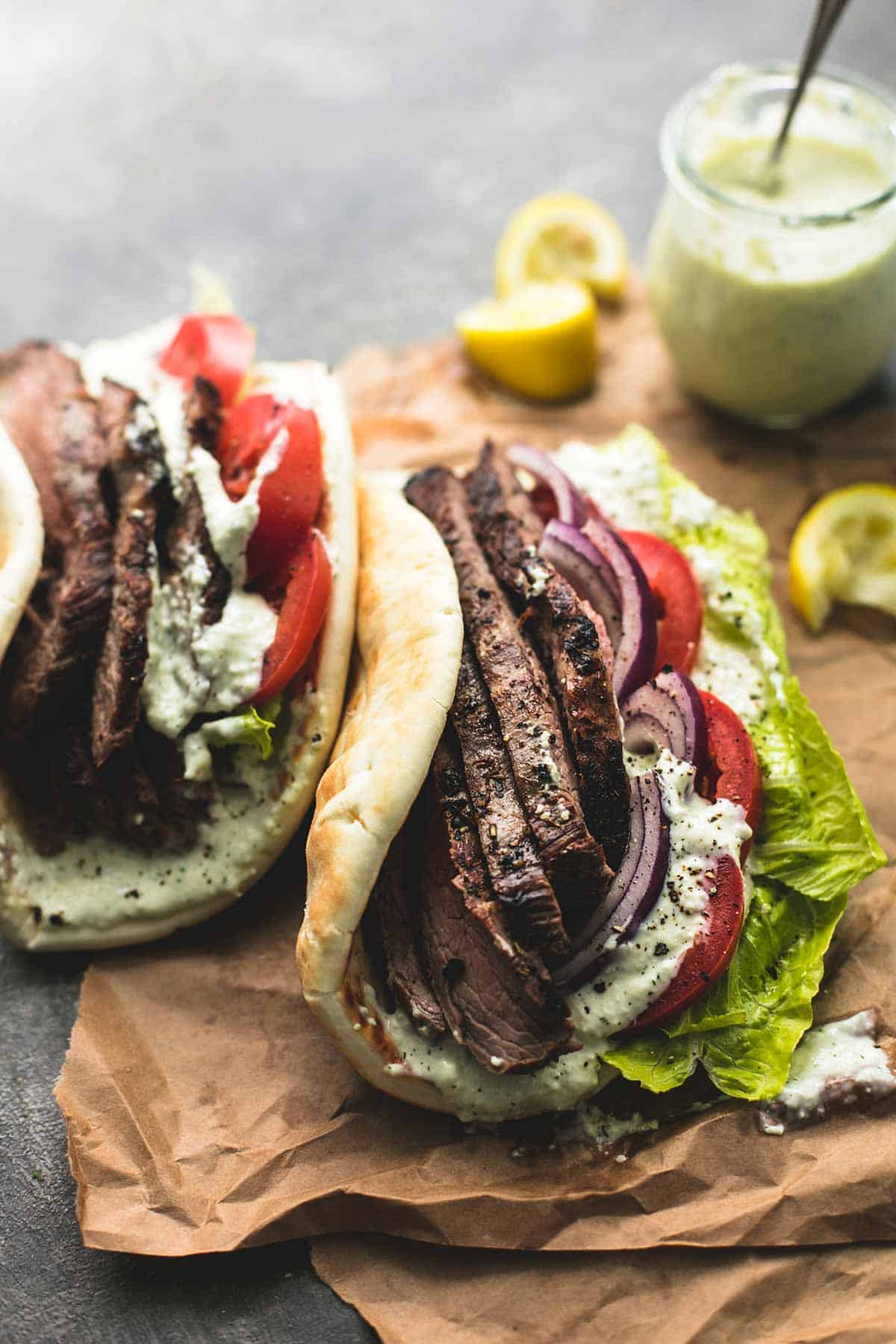 steak gyros with tzatziki cucumber sauce with more sauce in a jar and squeezed lemon slices in the background.