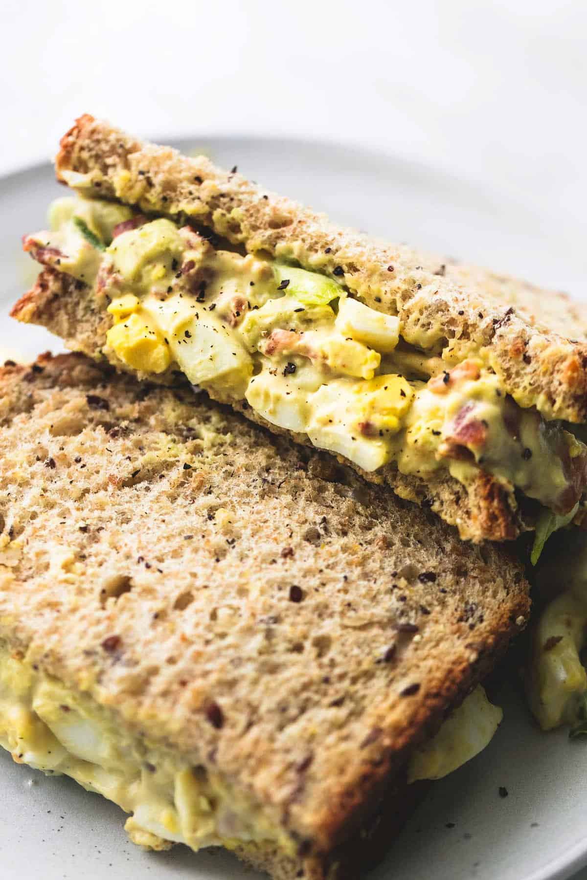close up of a bacon avocado egg salad sandwich cut in half on a plate.