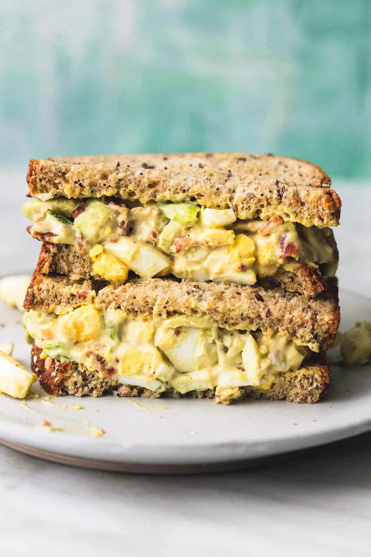 two bacon avocado egg salad sandwich halves stacked on top of each other on a plate.