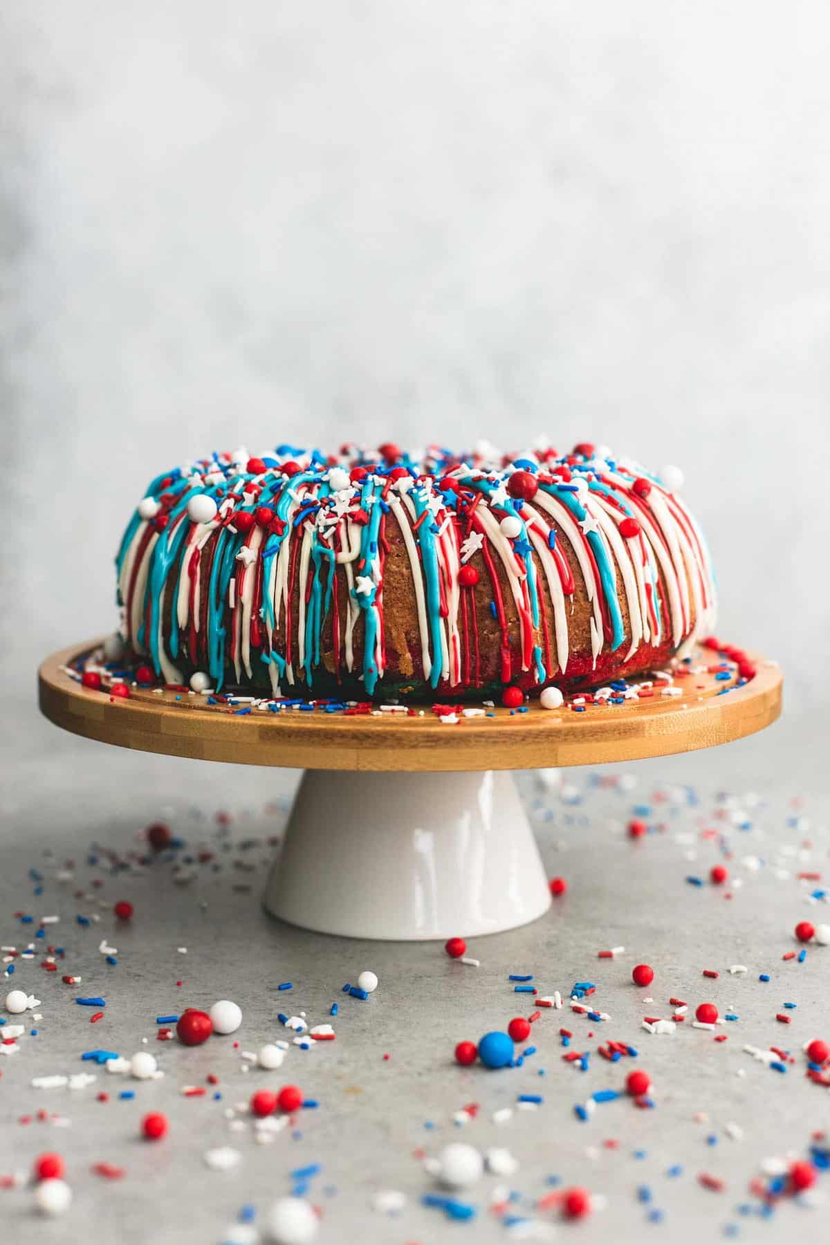 firecracker bundt cake frosted with red, white, and blue frosting and topped with sprinkles on a cake surrounded by more sprinkles.