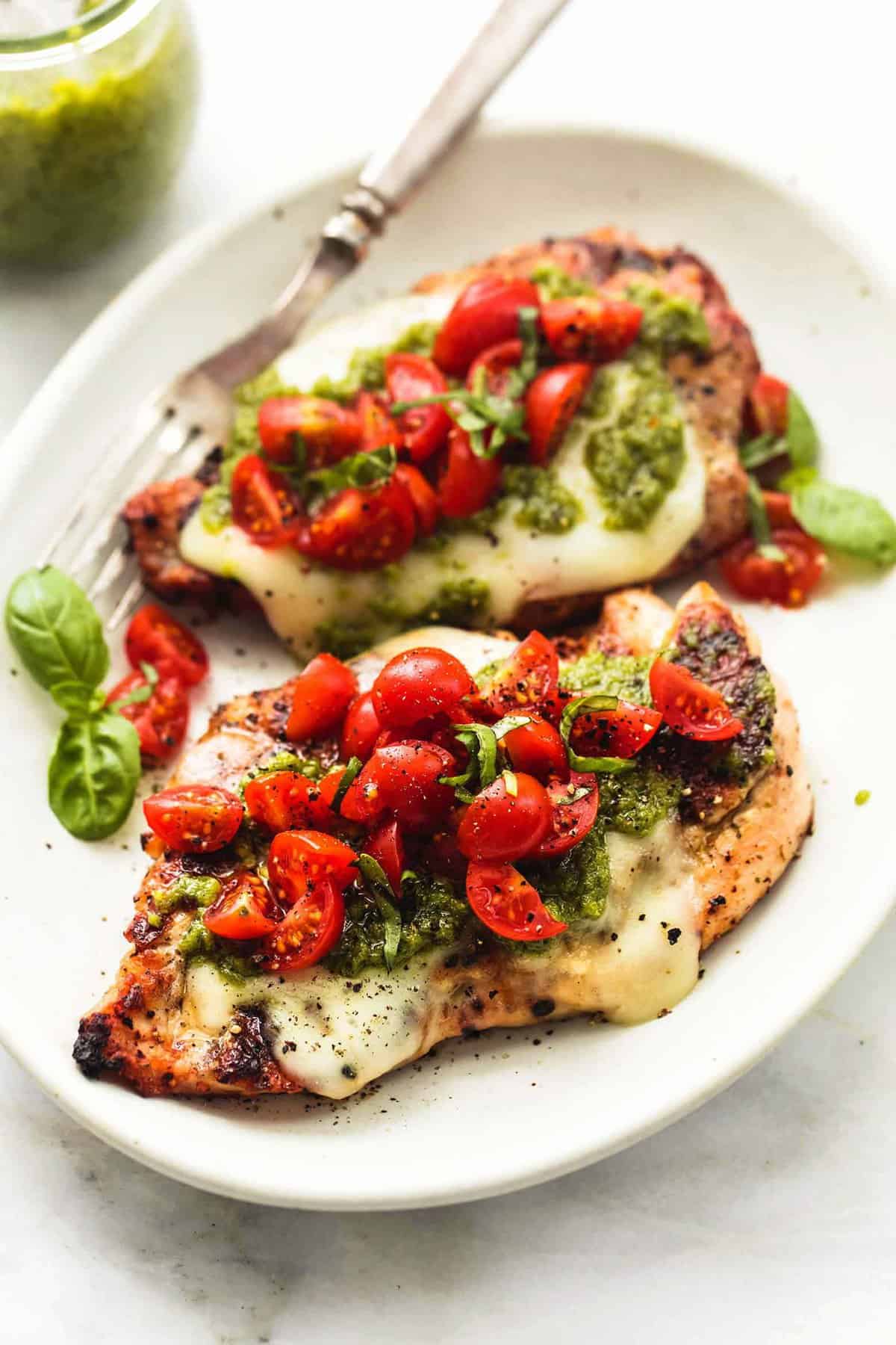 two grilled chicken breasts on plate topped with cheese, pesto, and tomatoes