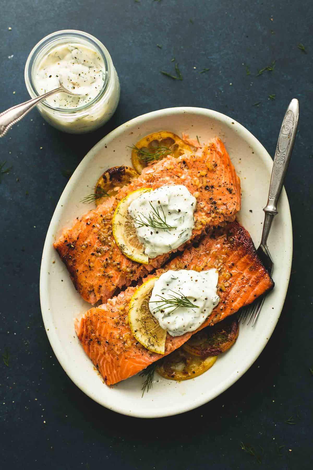 top view of baked salmon with creamy lemon dill sauce with a fork on a plate with a jar of more sauce with a spoon on the side.