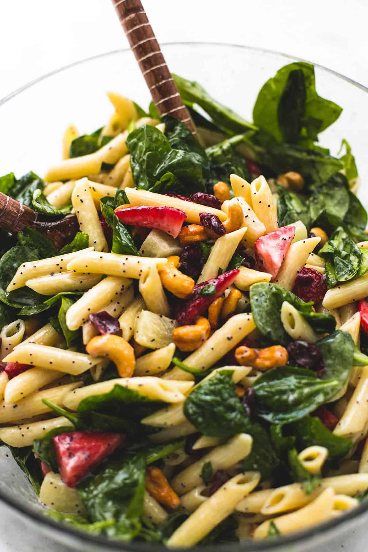 close up of strawberry spinach pasta salad with orange poppy seed dressing with two wooden serving spoons in a glass bowl.