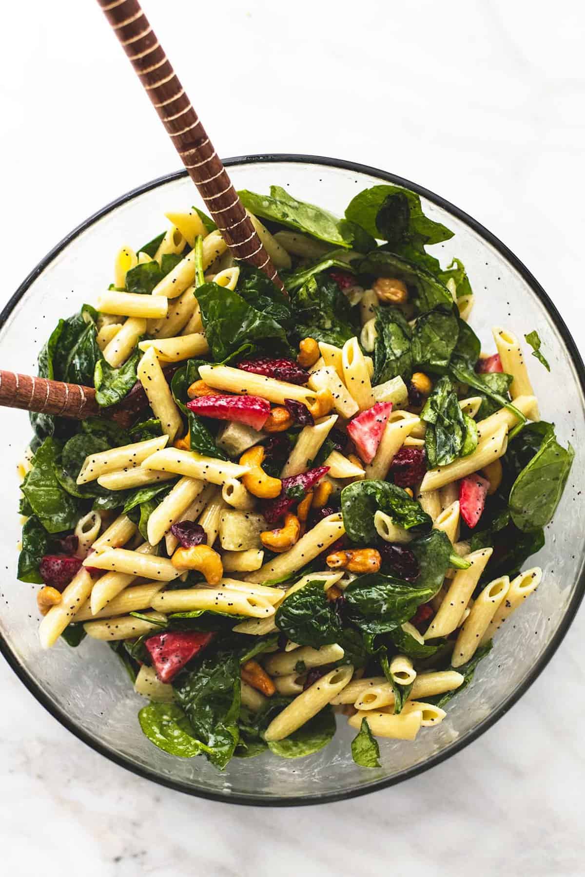 top view of strawberry spinach pasta salad with orange poppy seed dressing with two wooden serving spoons in a glass bowl.