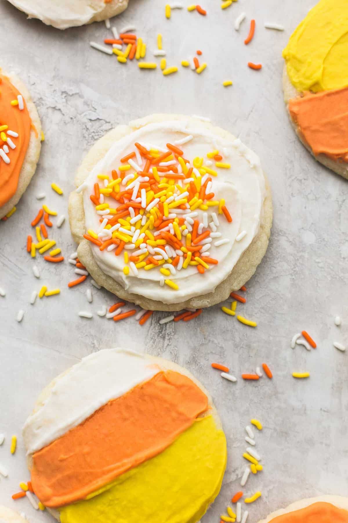 close up top view of a white frosted candy corn sugar cookie with sprinkles on top with more cookies frosted differently around it.