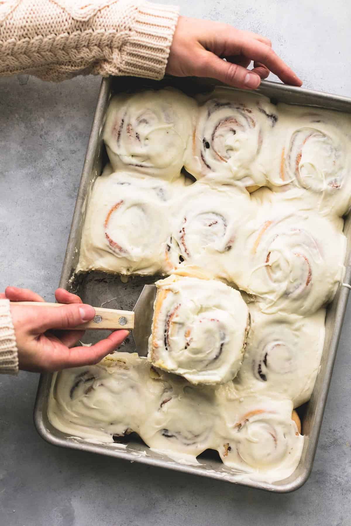 top view of a hand scooping up a eggnog cinnamon roll with a spatula from a baking sheet of eggnog cinnamon rolls.